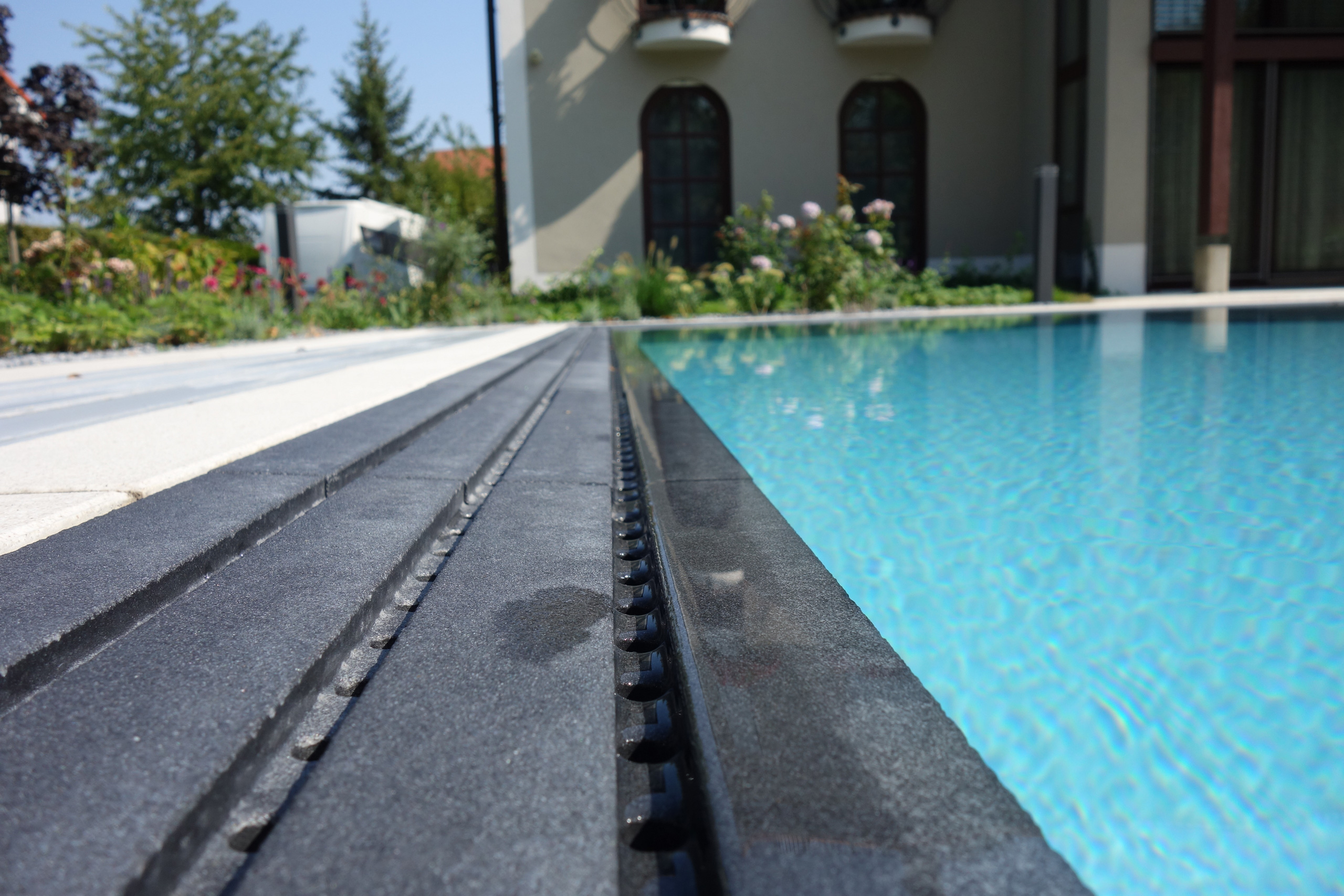 Detailansicht des Überlaufbeckens mit Überlaufrinne und Gitterrost aus dunklem Stein, Wasserfläche im Hintergrund