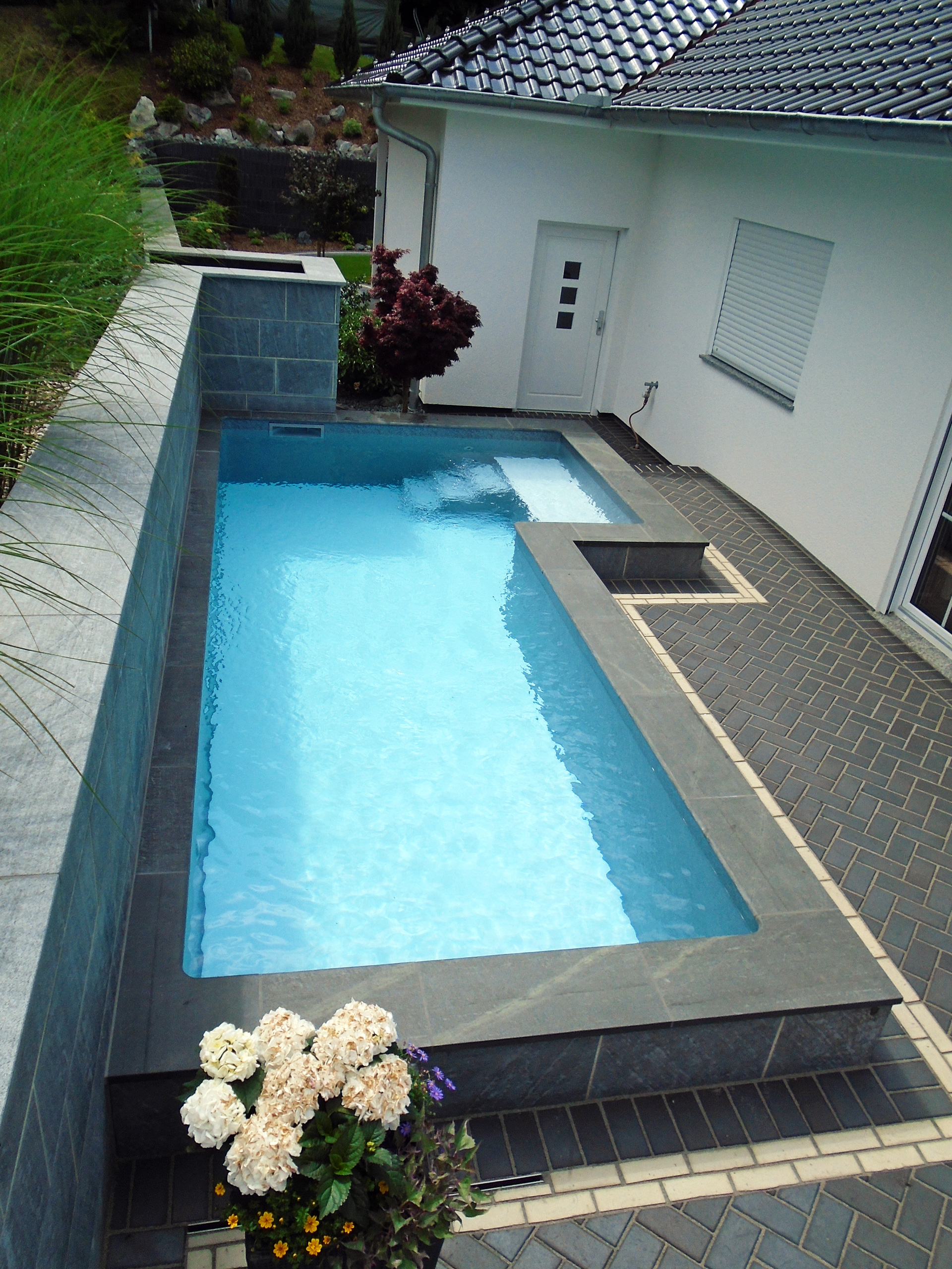 Blick auf das L-förmige Schwimmbecken mit Treppe und Podest, umgeben von dunklen Fliesen