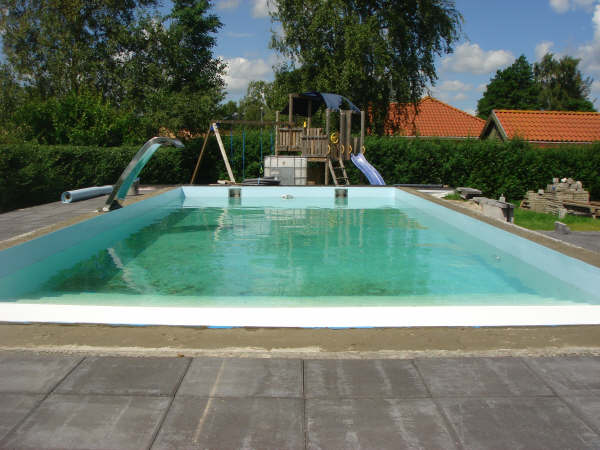 Rechteckiges Becken mit GSA und Schwalldusche, umgeben von einer Hecke und einem Kinderspielplatz.