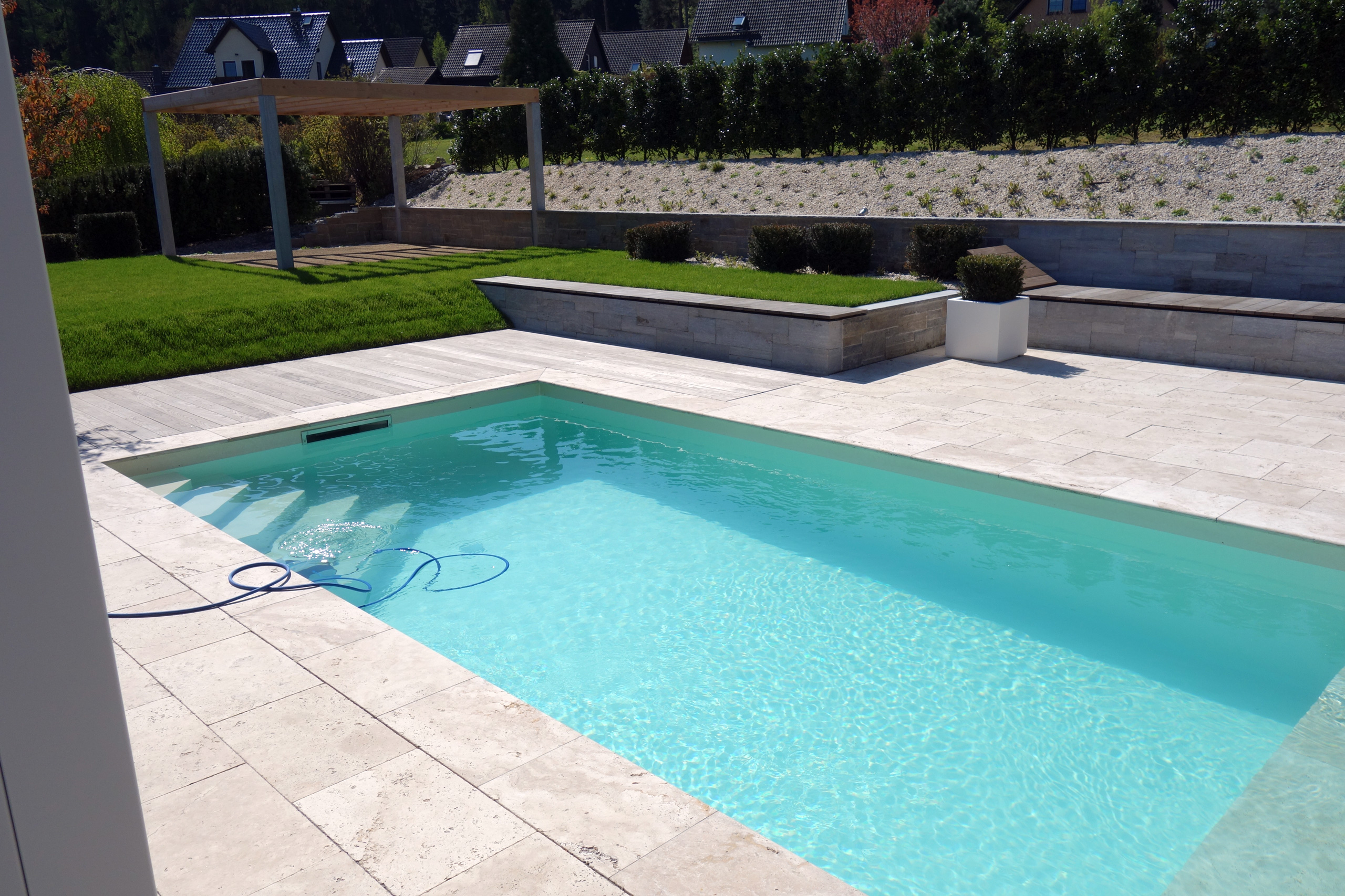 Schwimmbecken mit Strömungsturbine und Rollladenabdeckung, umgeben von heller Terrasse und Garten.