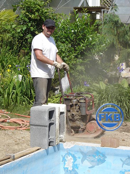Mann verdichtet den Untergrund neben einem hellblauen Becken mit Fischmotiven und vorbereiteten Hohlblocksteinen.