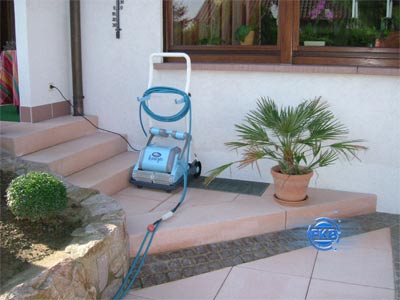 Poolroboter mit Kabel und Schlauch auf einer Terrasse neben einer Palme und Treppen
