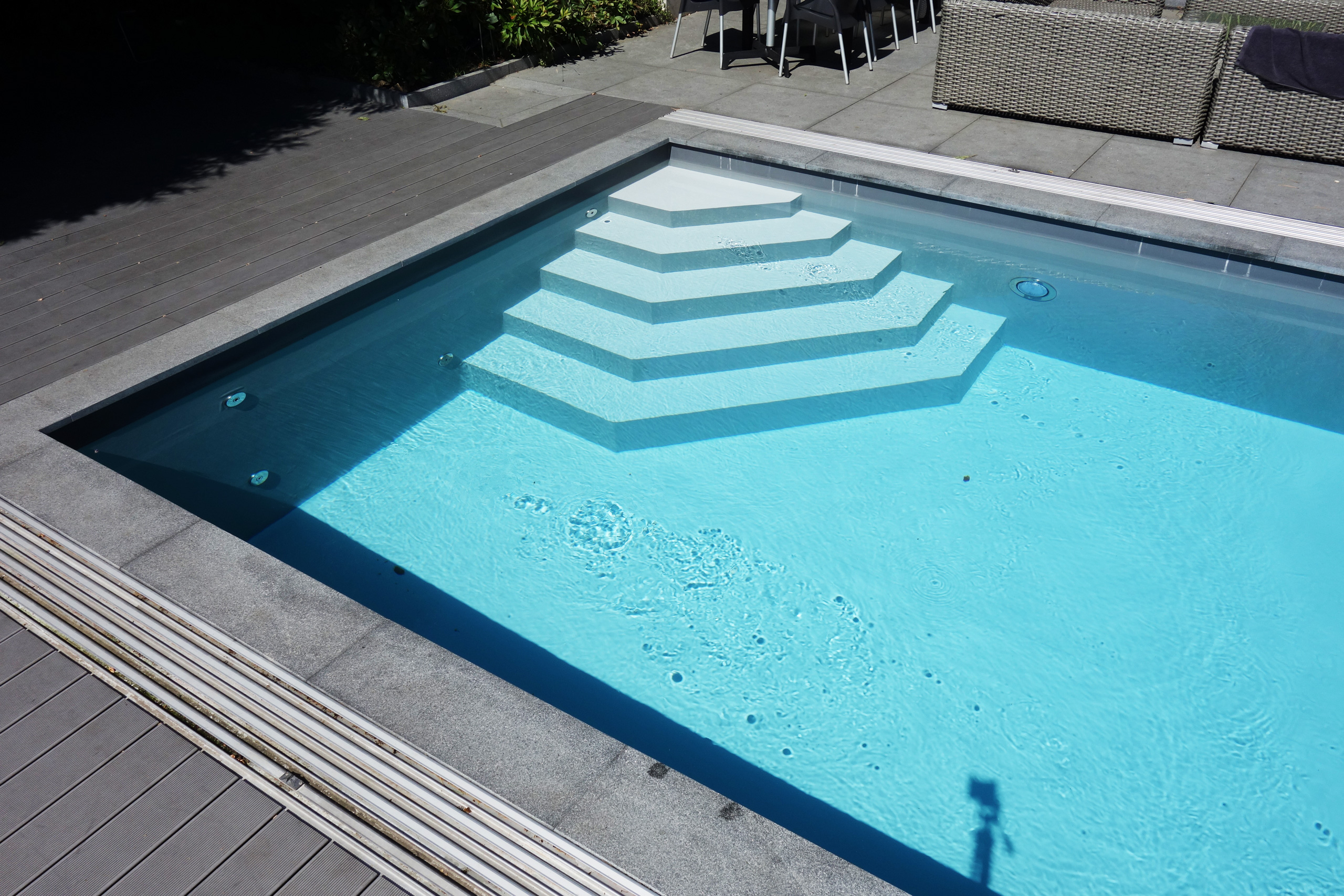 Detailansicht des Betonbeckens mit weißer Ecktreppe und Sitzbank, umgeben von Holzterrasse und Steinplatten.
