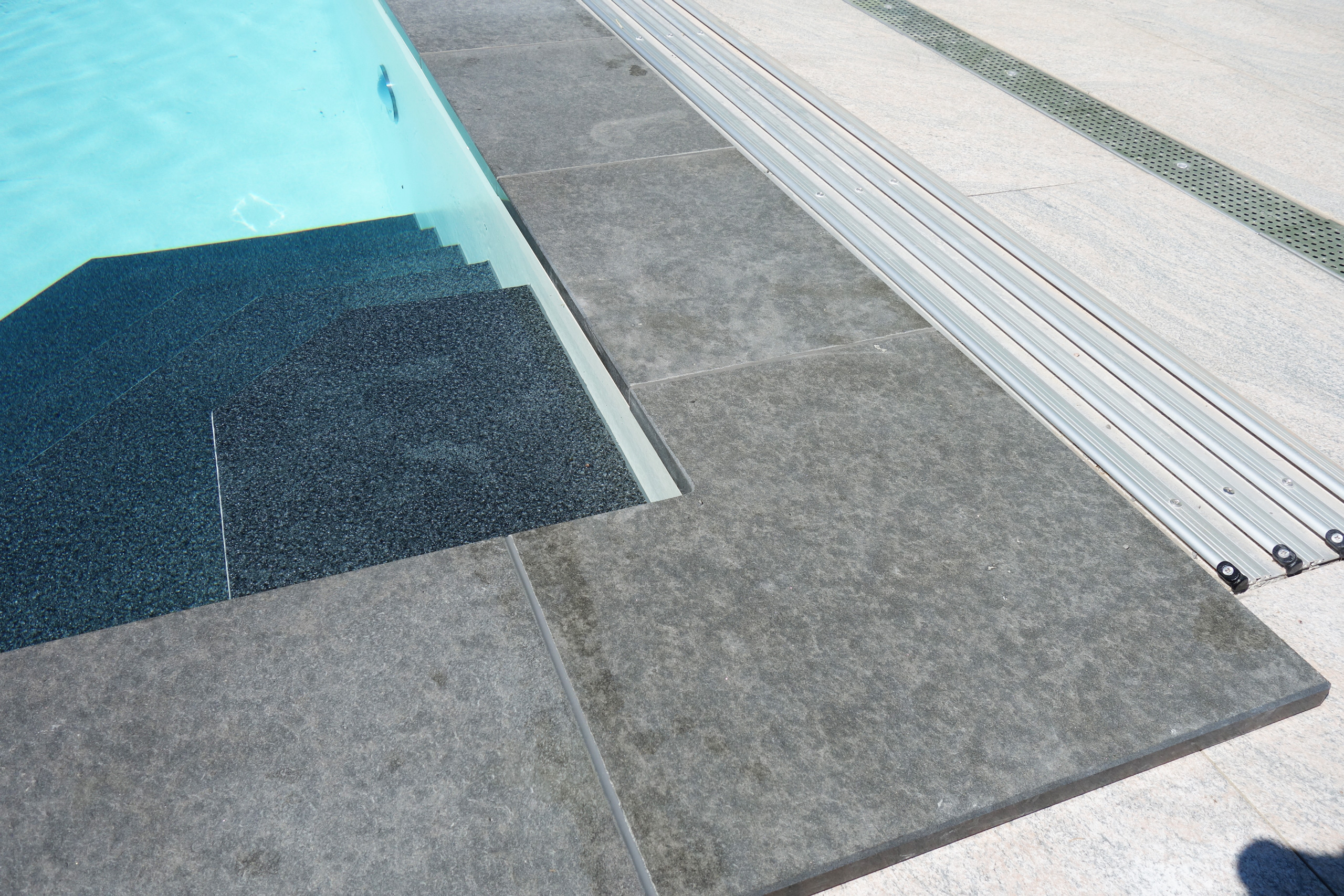 Pooltreppe mit dunkler Folie und hellblauer Wasseroberfläche, umgeben von grauen Steinplatten und Rolladenabdeckung