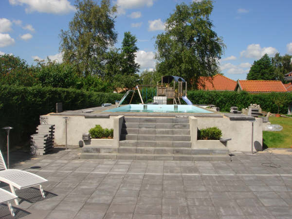 Teilansicht eines eingelassenen Pools mit Treppenaufgang, umgeben von einer gepflasterten Terrasse und Hecken.