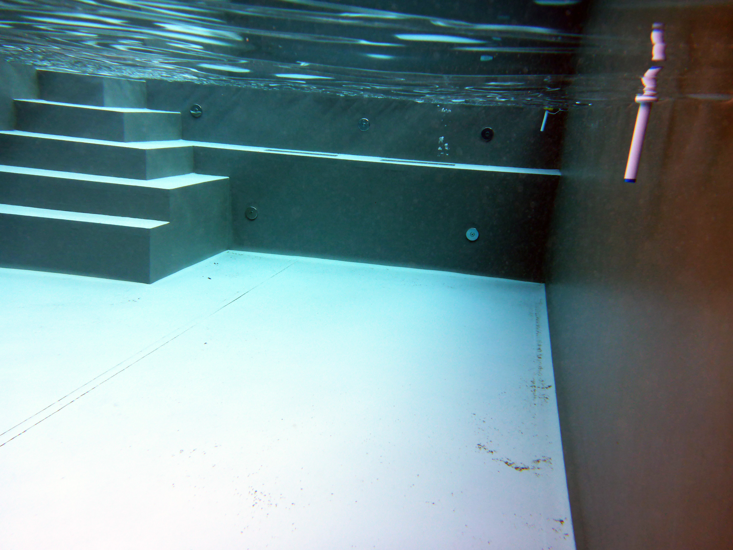 Unterwasseransicht eines Betonbeckens mit Ecktreppe und Podest, die Wasseroberfläche spiegelt sich.