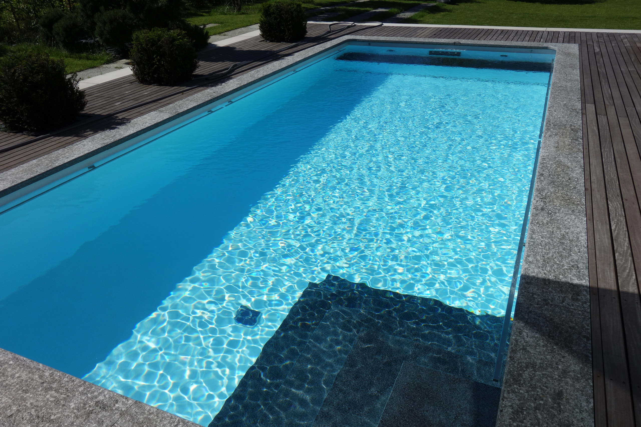 Rechteckbecken mit blauer Folie, dunkler Ecktreppe und Podest, umgeben von Holzterrasse und Rasen