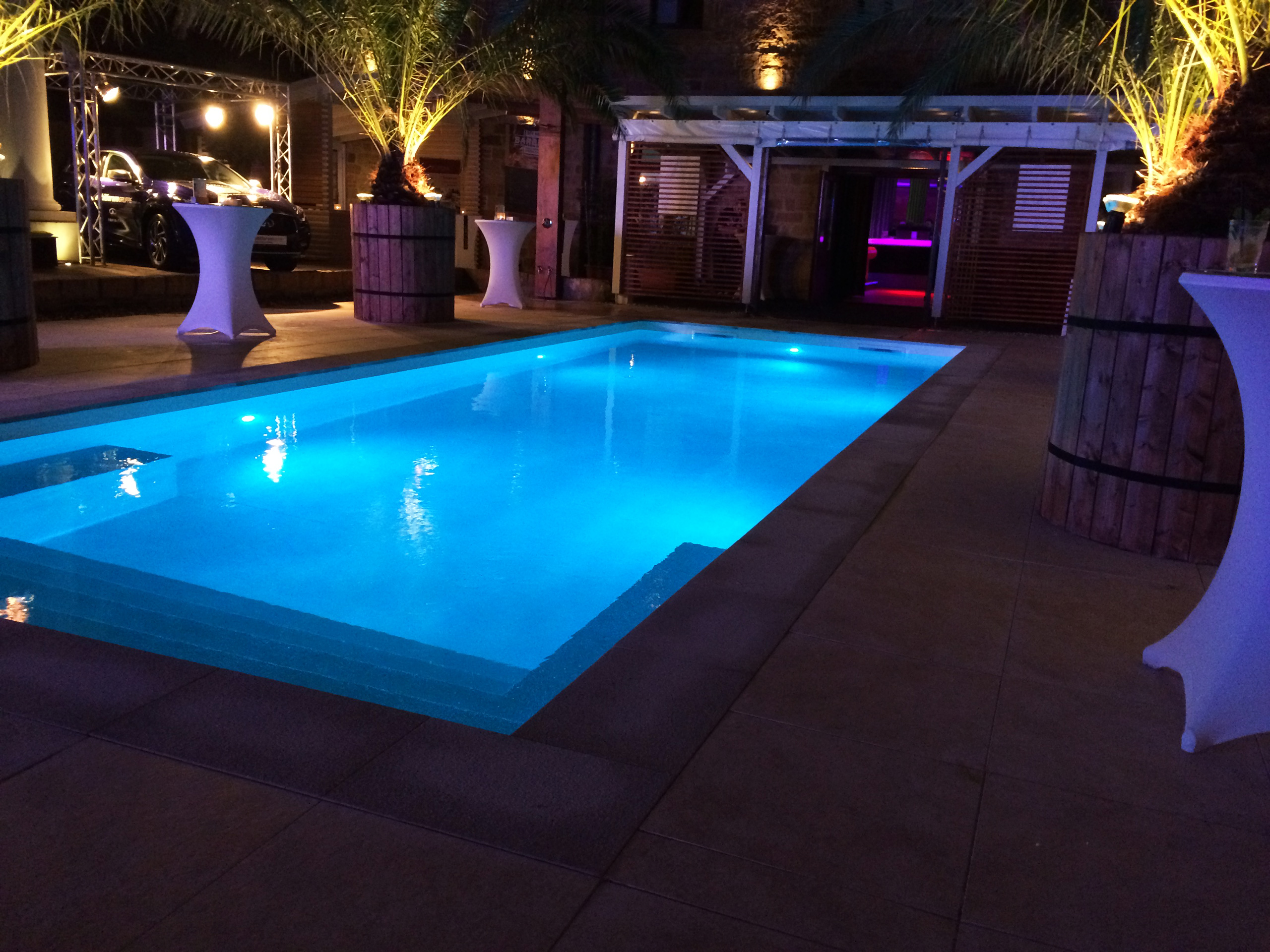 Beleuchtetes Schwimmbad mit gerader Treppe und Podest bei Nacht, umgeben von Palmen und Event-Dekoration.