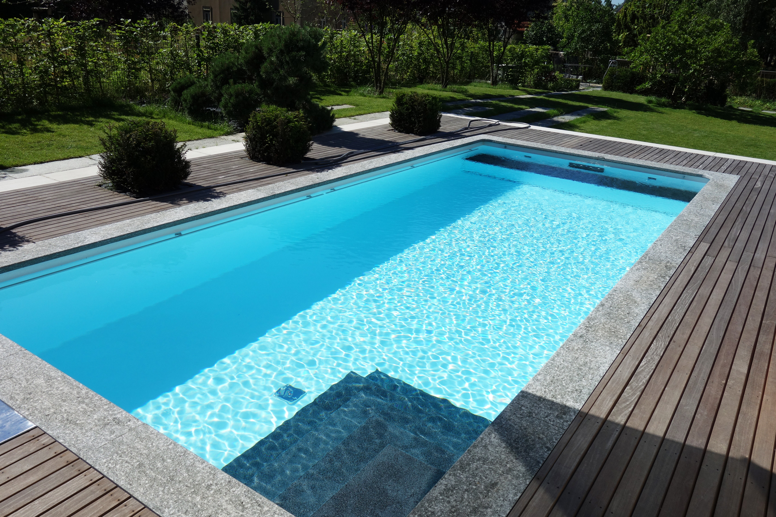 Rechteckbecken mit blauer Folie, Ecktreppe und Unterflurabdeckung, umgeben von Holzdeck und Grünfläche.
