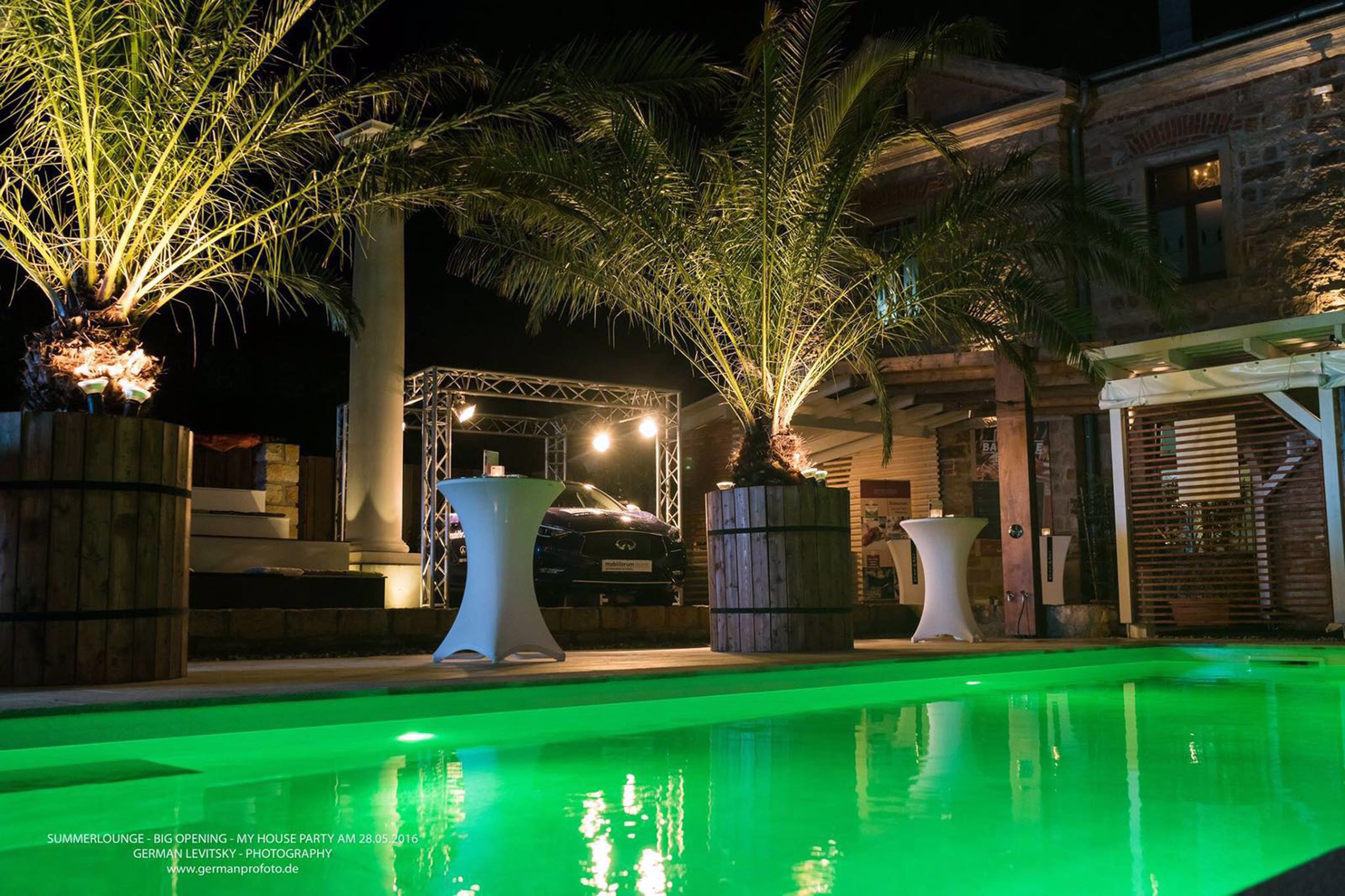 Grün beleuchtetes Schwimmbad bei Nacht, umgeben von Palmen und Steingebäuden, mit Stehtischen am Beckenrand.