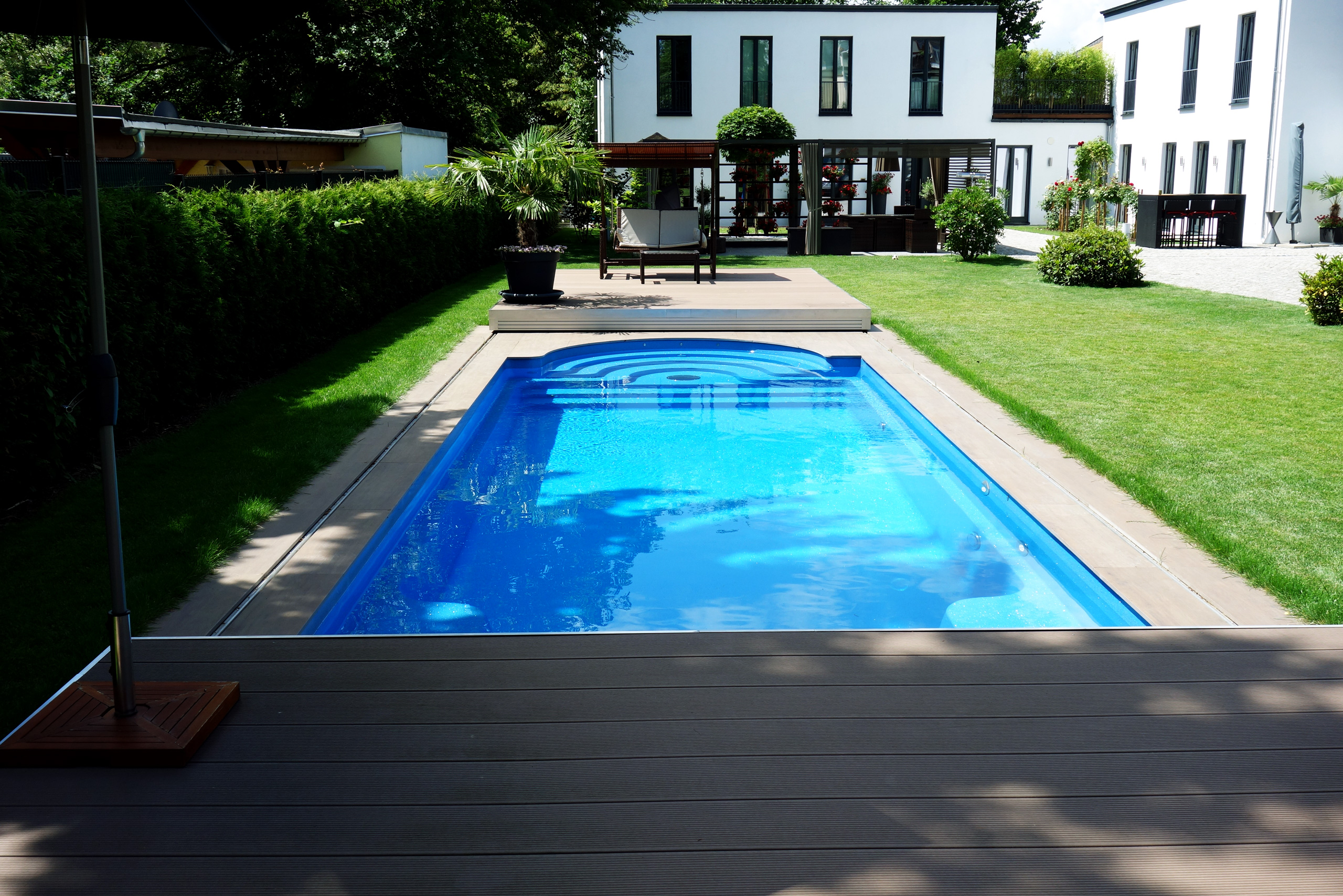 Blick auf das blaue Compass Pool Schwimmbecken Java 101 mit Terrassenabdeckung und Gartenanlage