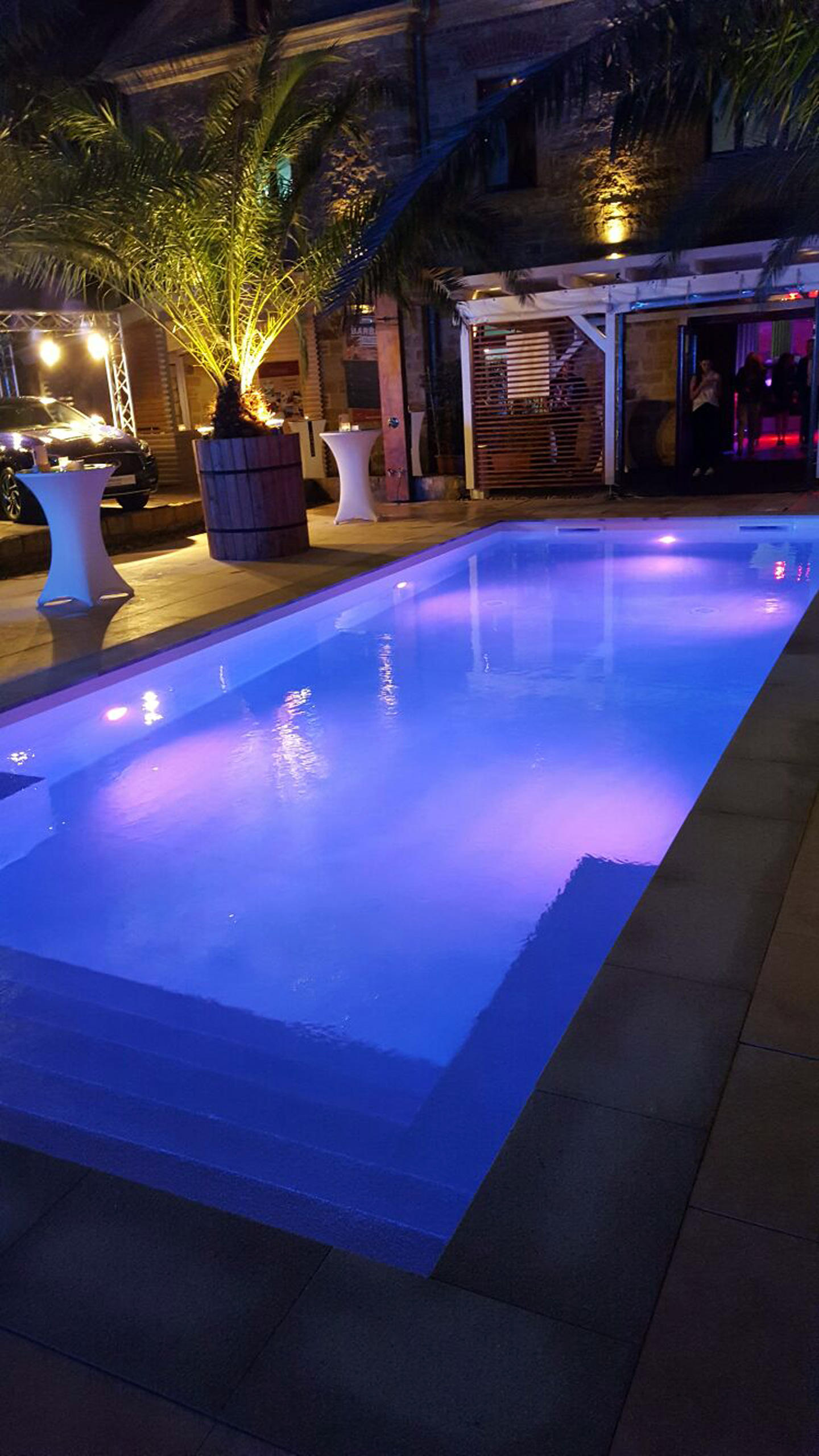 Beleuchtetes Schwimmbad mit gerader Treppe und Podest bei Nacht, umgeben von Palmen und Event-Dekoration.