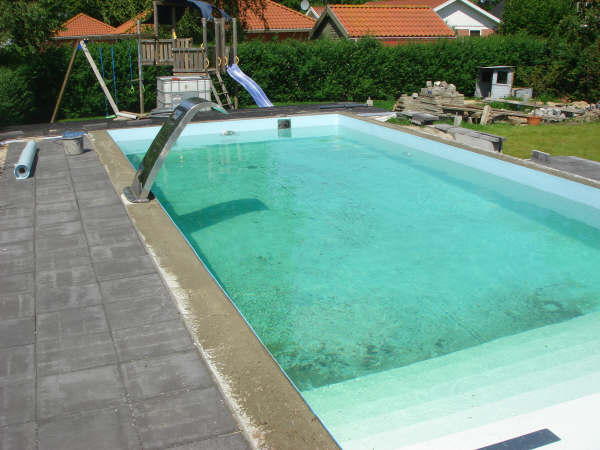 Edelstahl-Schwalldusche am Beckenrand eines Pools mit Treppe und GSA, umgeben von Pflastersteinen.