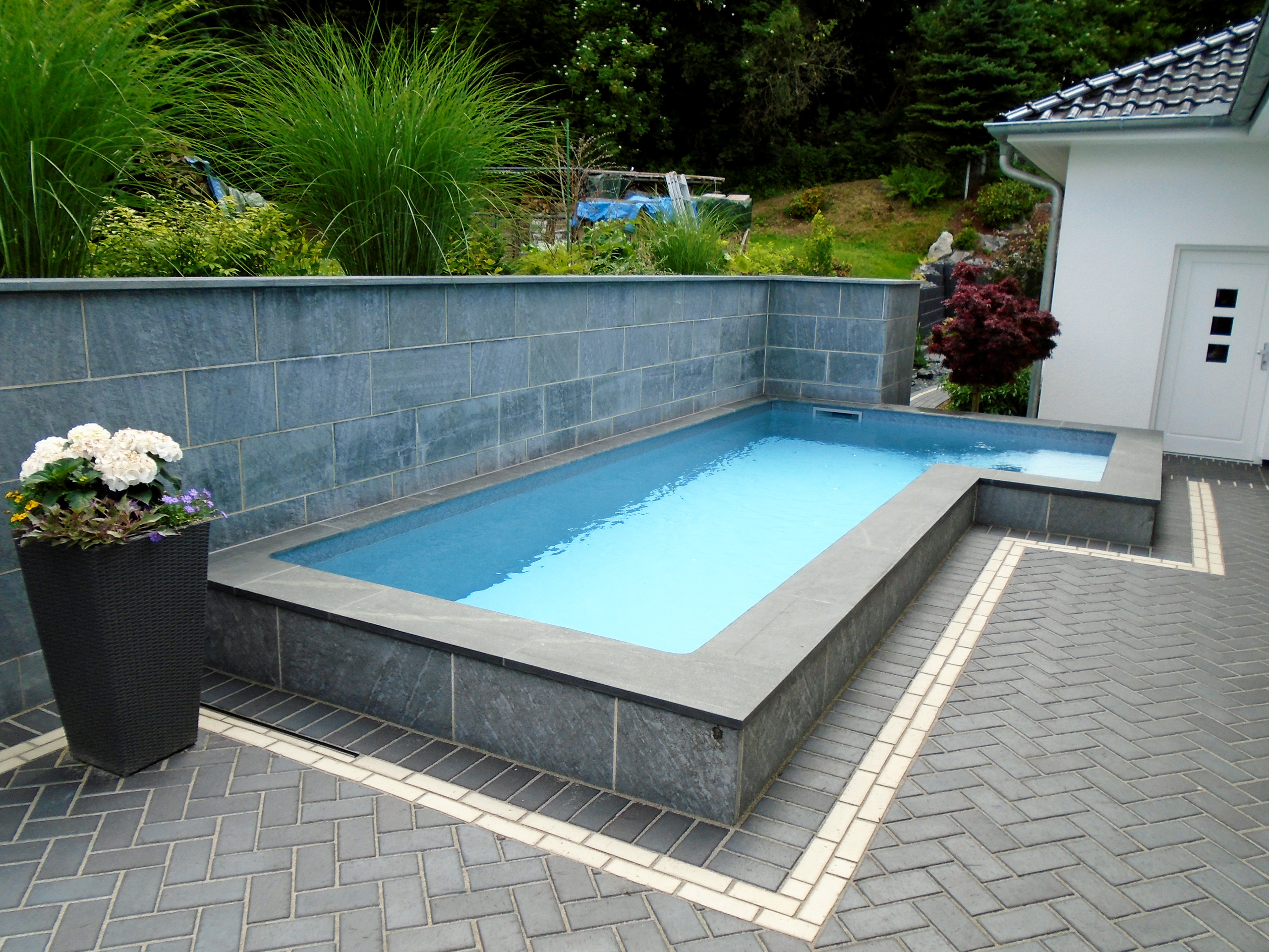 L-Form Schwimmbecken mit Treppe und Podest, umrandet von dunklen Fliesen und Pflastersteinen
