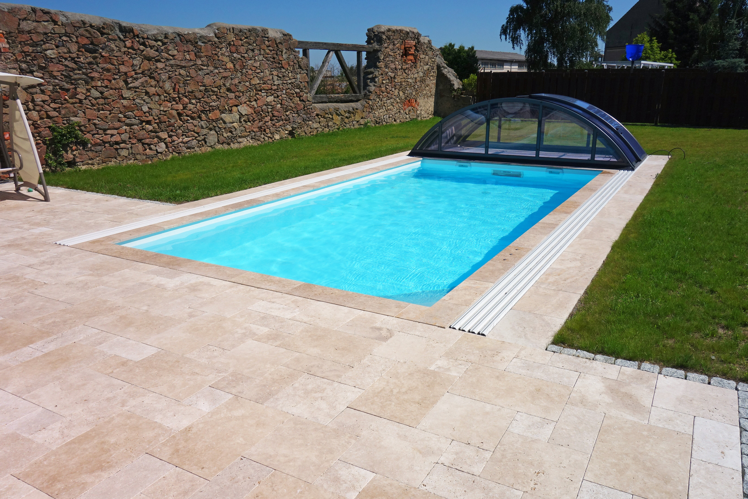 Schwimmbecken mit beigefarbenen Fliesen und einer halb geöffneten, transparenten Poolüberdachung im Garten.