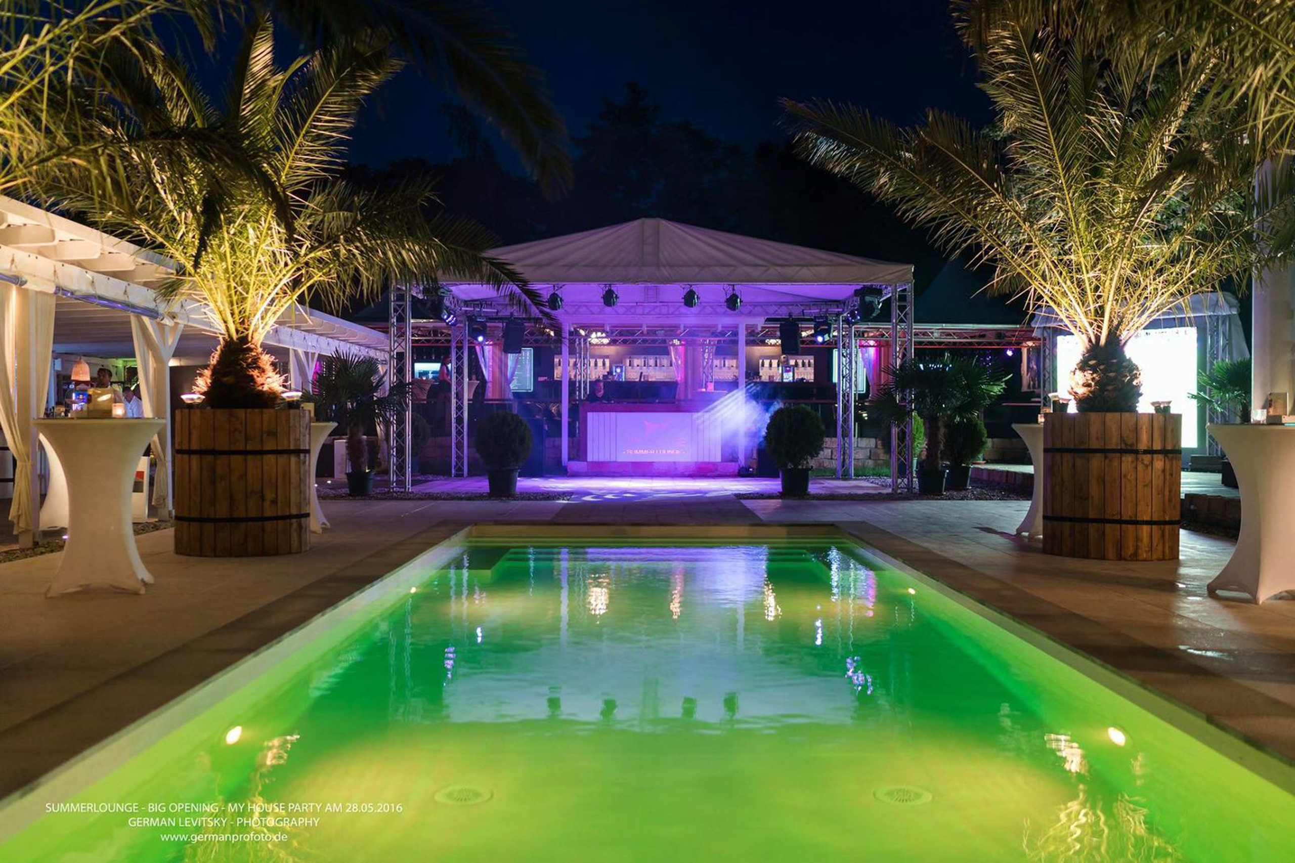 Schwimmbad mit grüner Beleuchtung bei Nacht, umgeben von Palmen und einer beleuchteten Bar