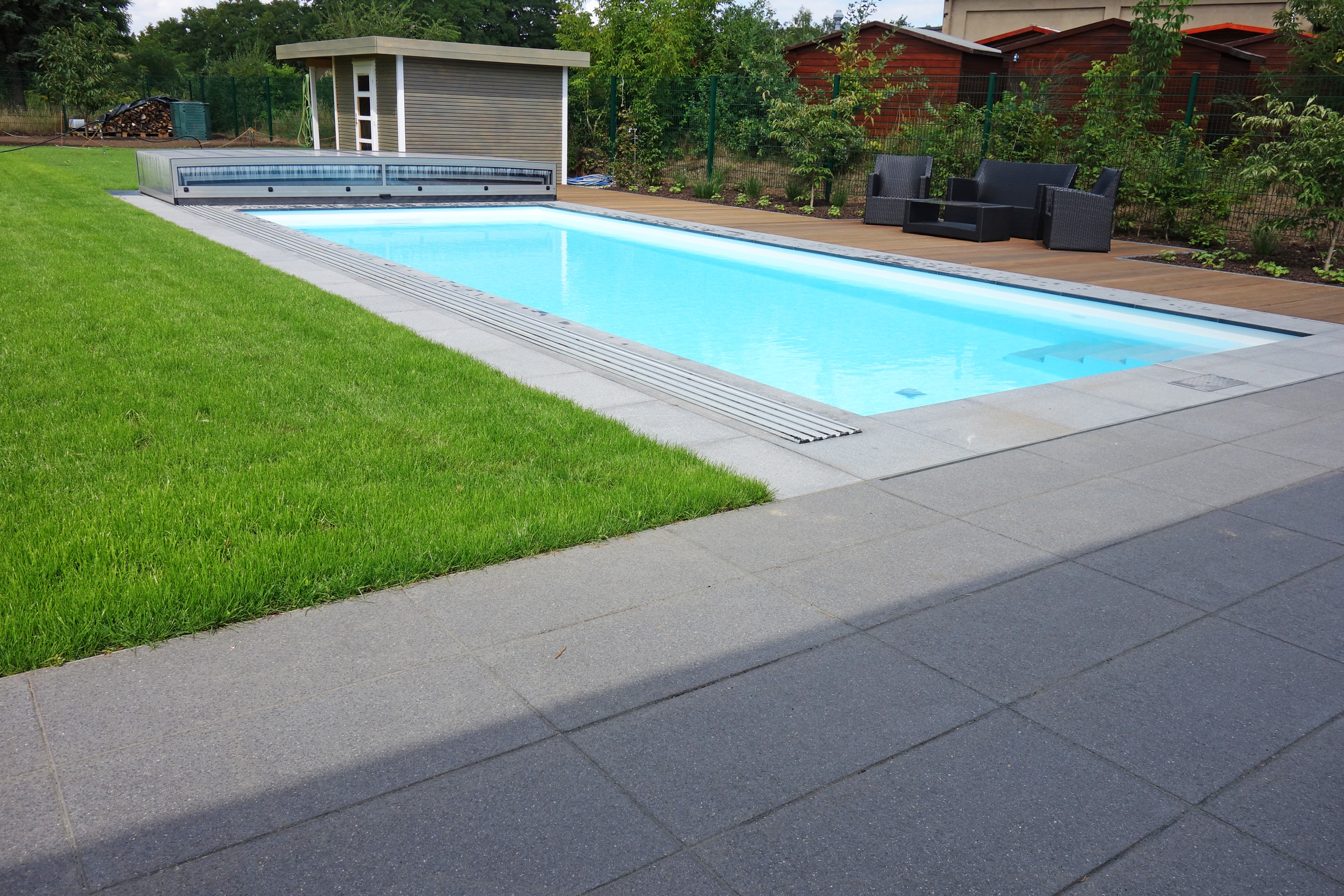 Rechteckiger Außenpool mit blauer Folie, grauen Fliesen und Holzterrasse, teilweise von Rasen umgeben