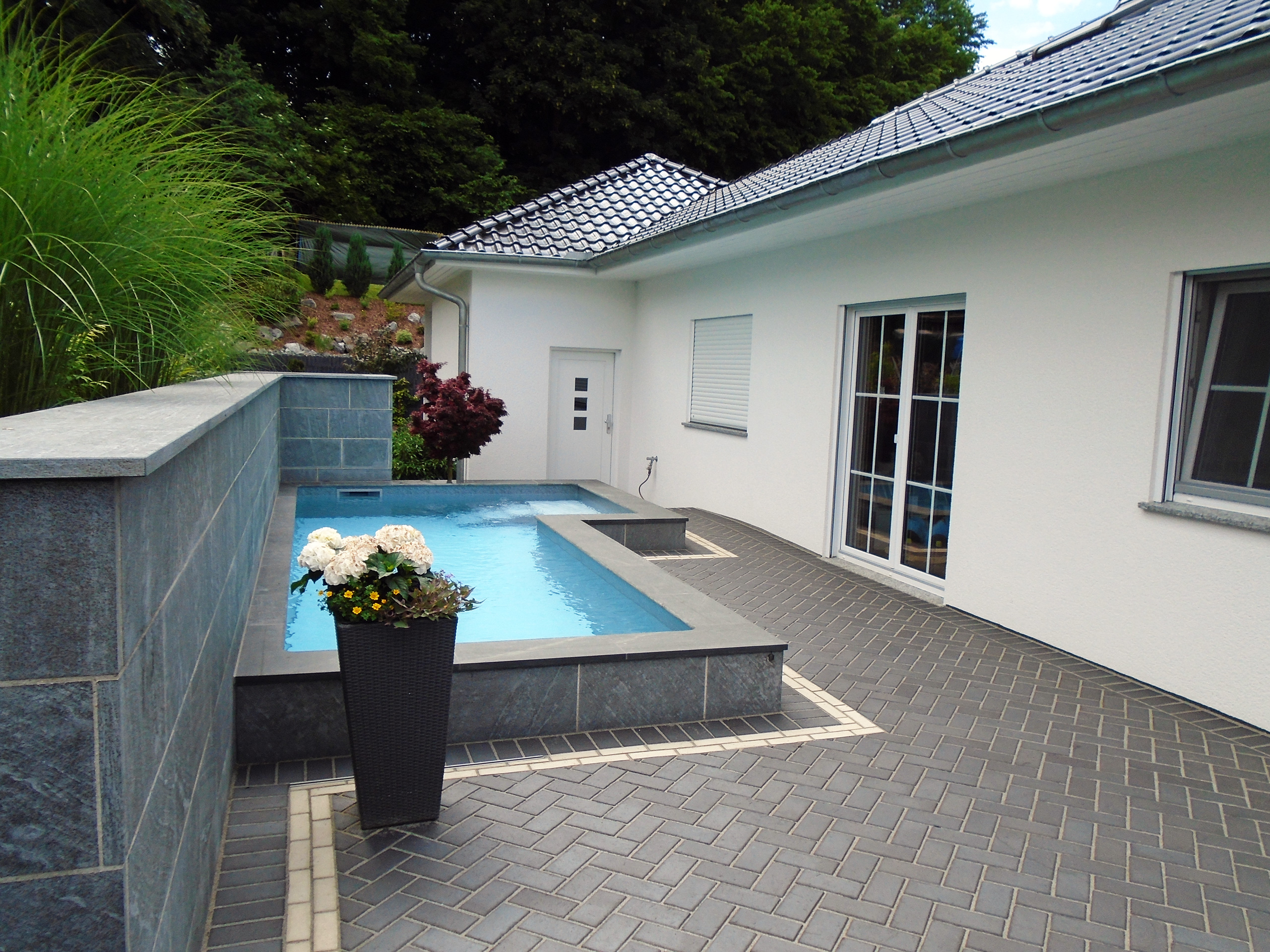 L-förmiges Schwimmbecken mit Treppe und Podest, umgeben von dunklen Steinplatten und einer weißen Hauswand