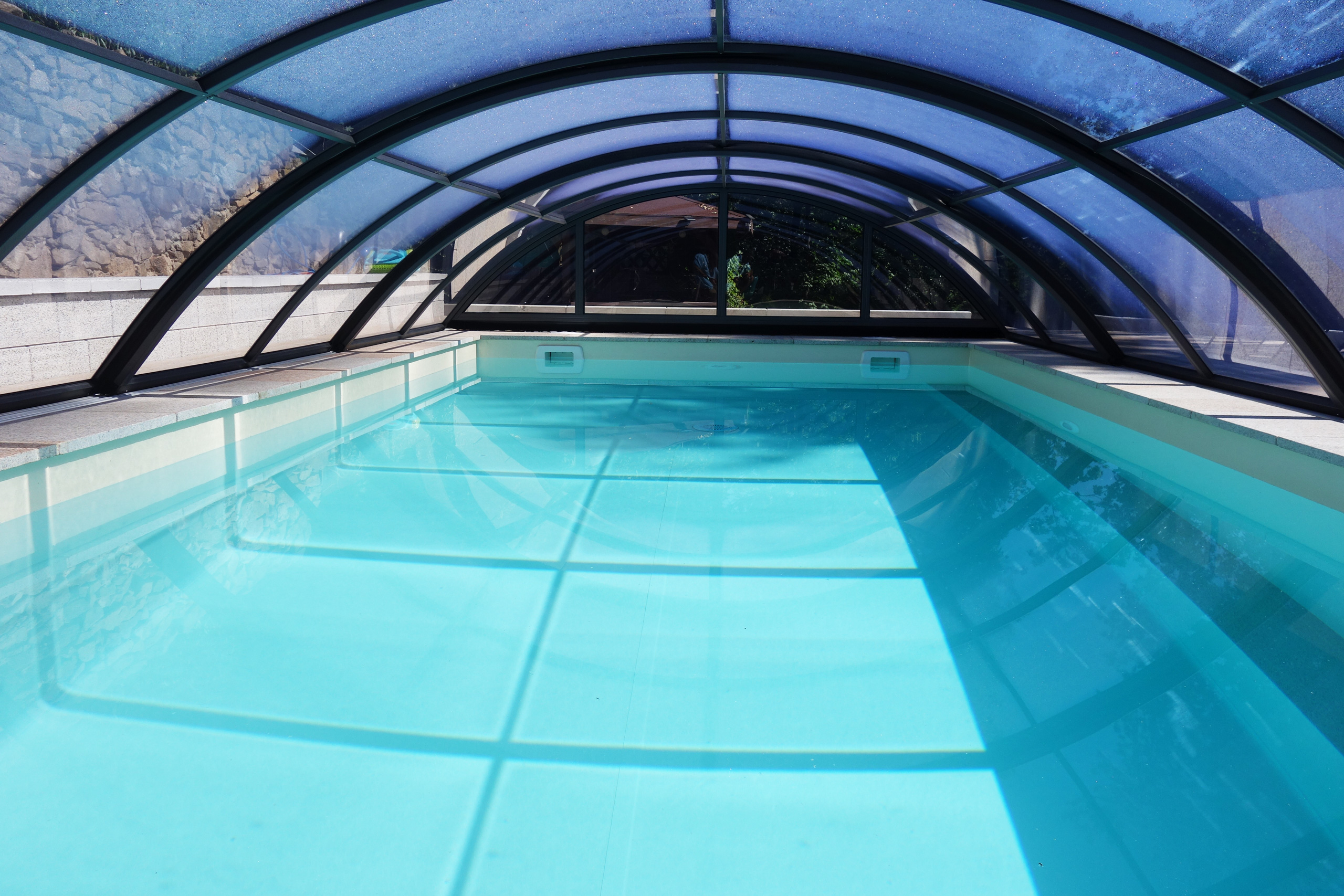 Blick in den Rechteckpool mit Ecktreppe und Stehstufe unter der transparenten Poolüberdachung Klasik Clear