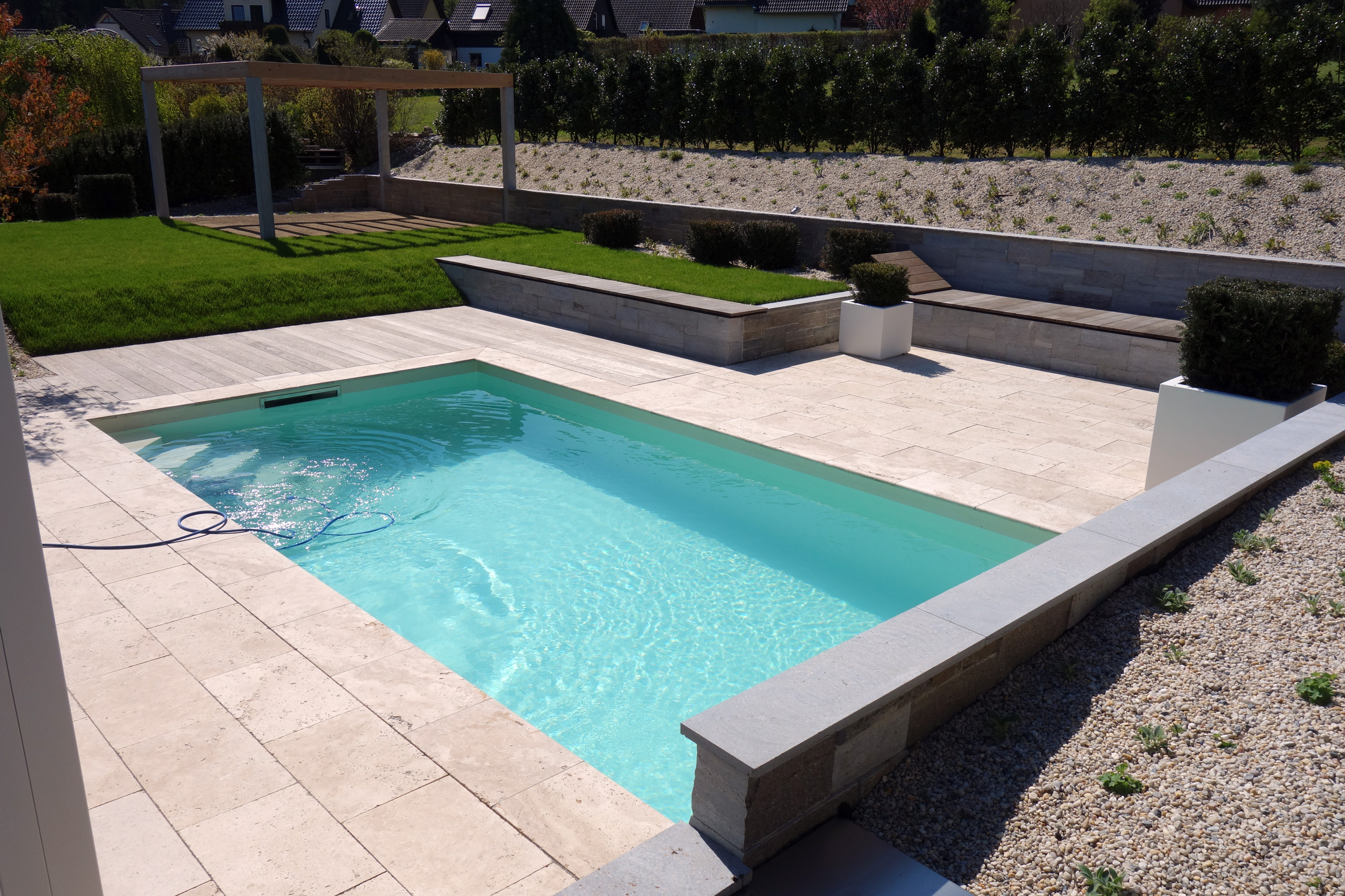 Schwimmbecken mit hellblauer Folie, Holzterrasse und beigefarbenen Steinplatten im Garten.