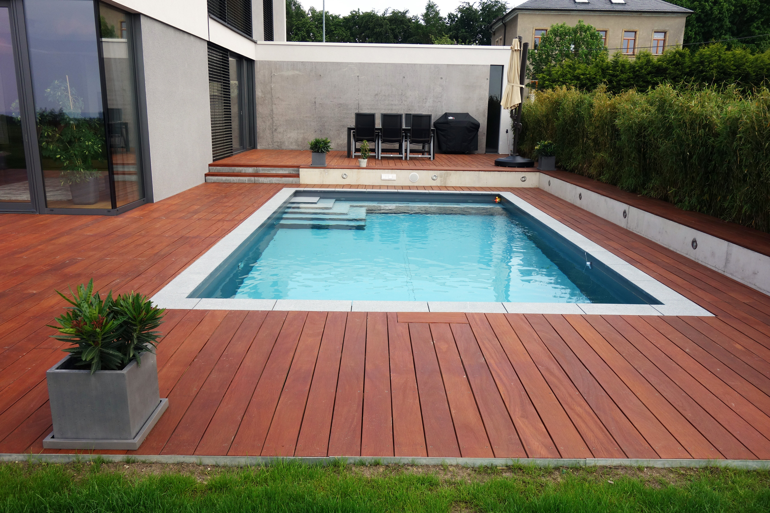 Gesamtaufnahme des Betonbeckens 3,5x7x1,5m mit Ecktreppe und Podest, umgeben von Holzterrasse und Garten.