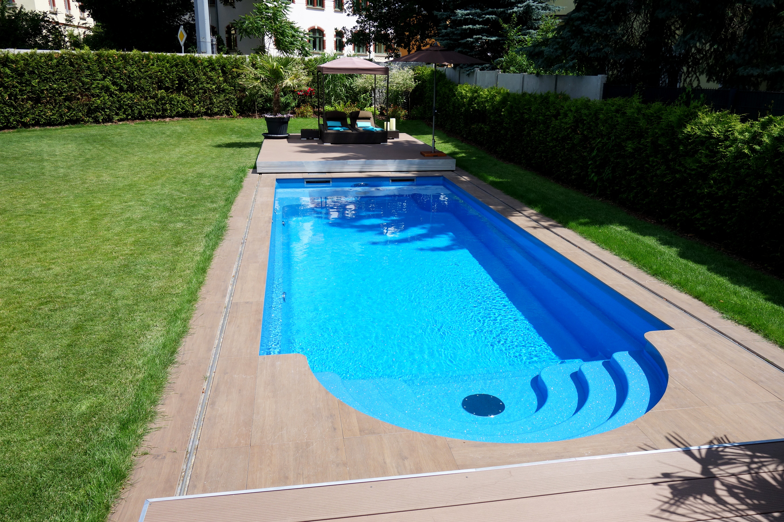 Blick auf das blaue Compass Pool Schwimmbecken Java 101 mit Terrassenabdeckung und Holzoptik-Umfassung.