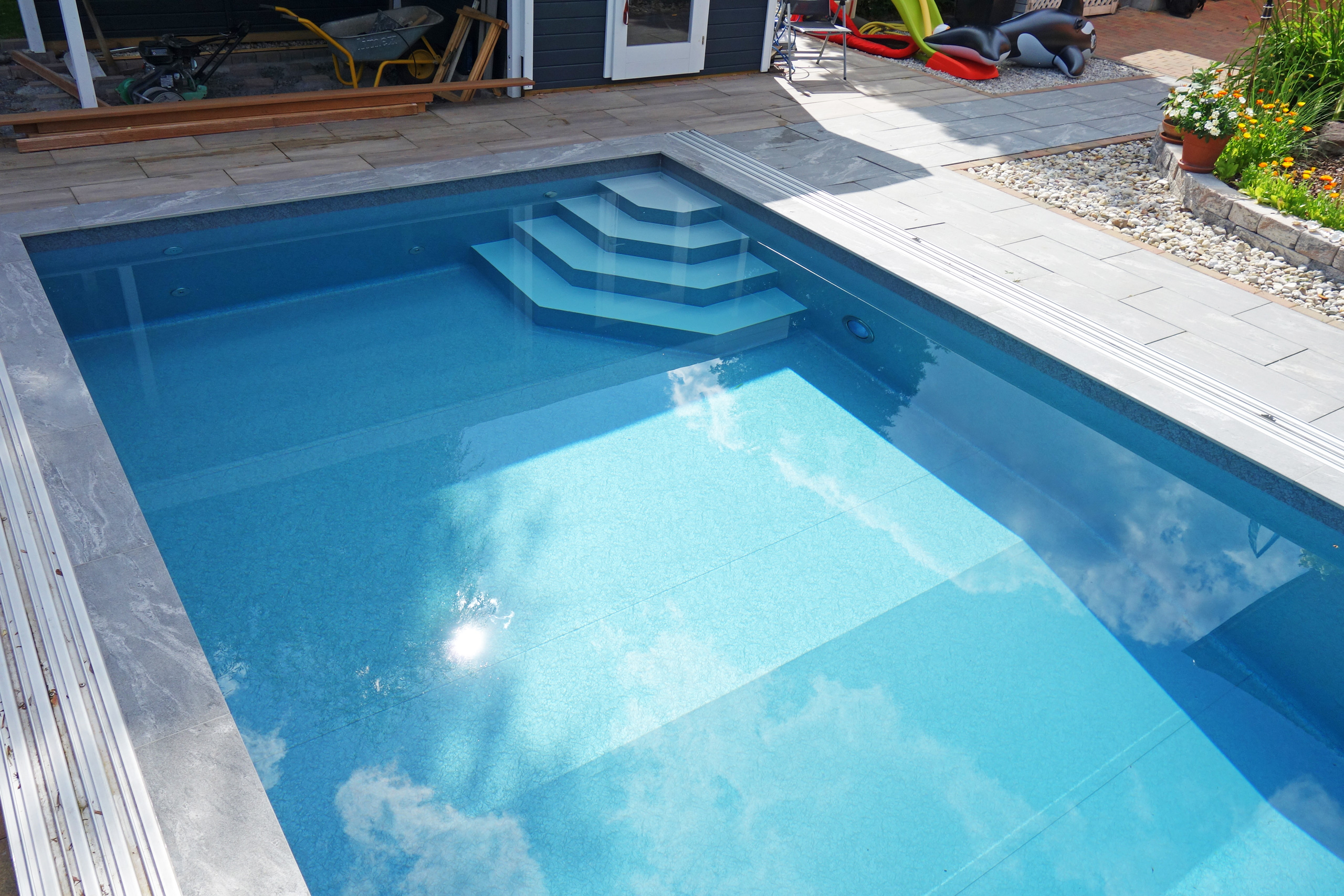 Blick auf das blaue Schwimmbecken mit Ecktreppe, Podest und geschlossener Überdachung