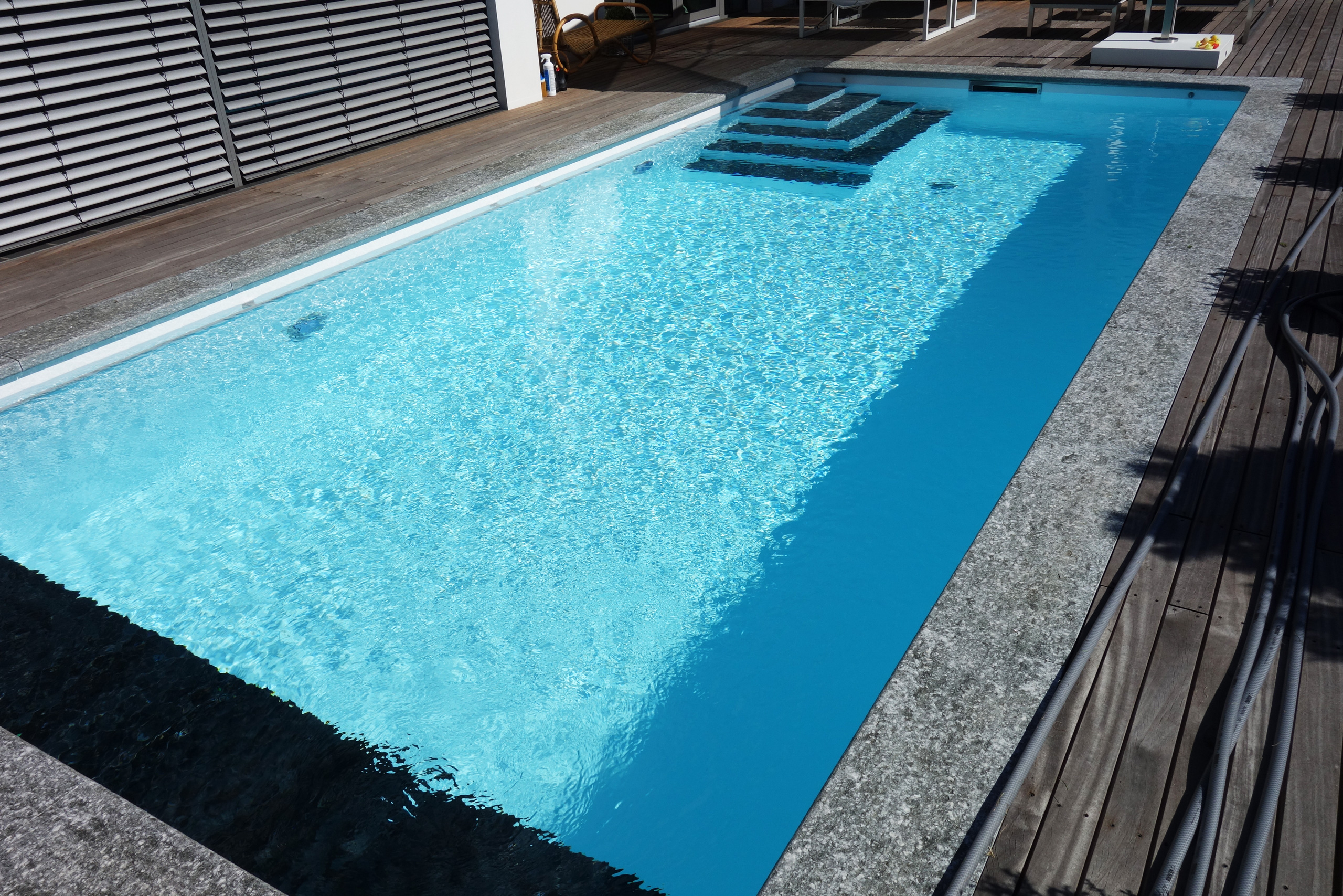Blick auf das Rechteckbecken mit blauer Folie, Ecktreppe und Holzterrasse mit Unterflurabdeckung.