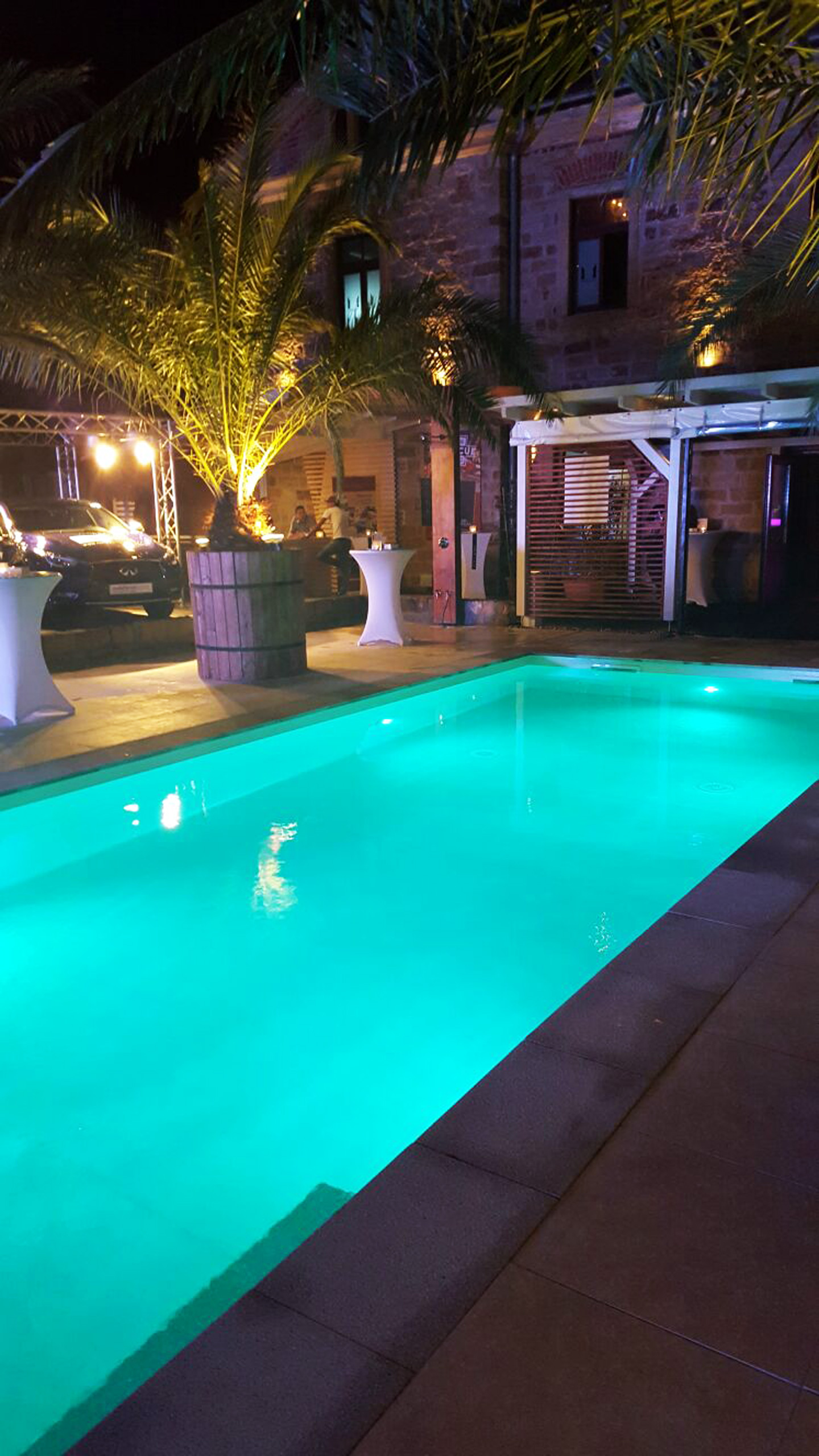 Beleuchtetes Schwimmbad mit gerader Treppe und Podest bei Nacht, umgeben von Palmen und einem Gebäude.