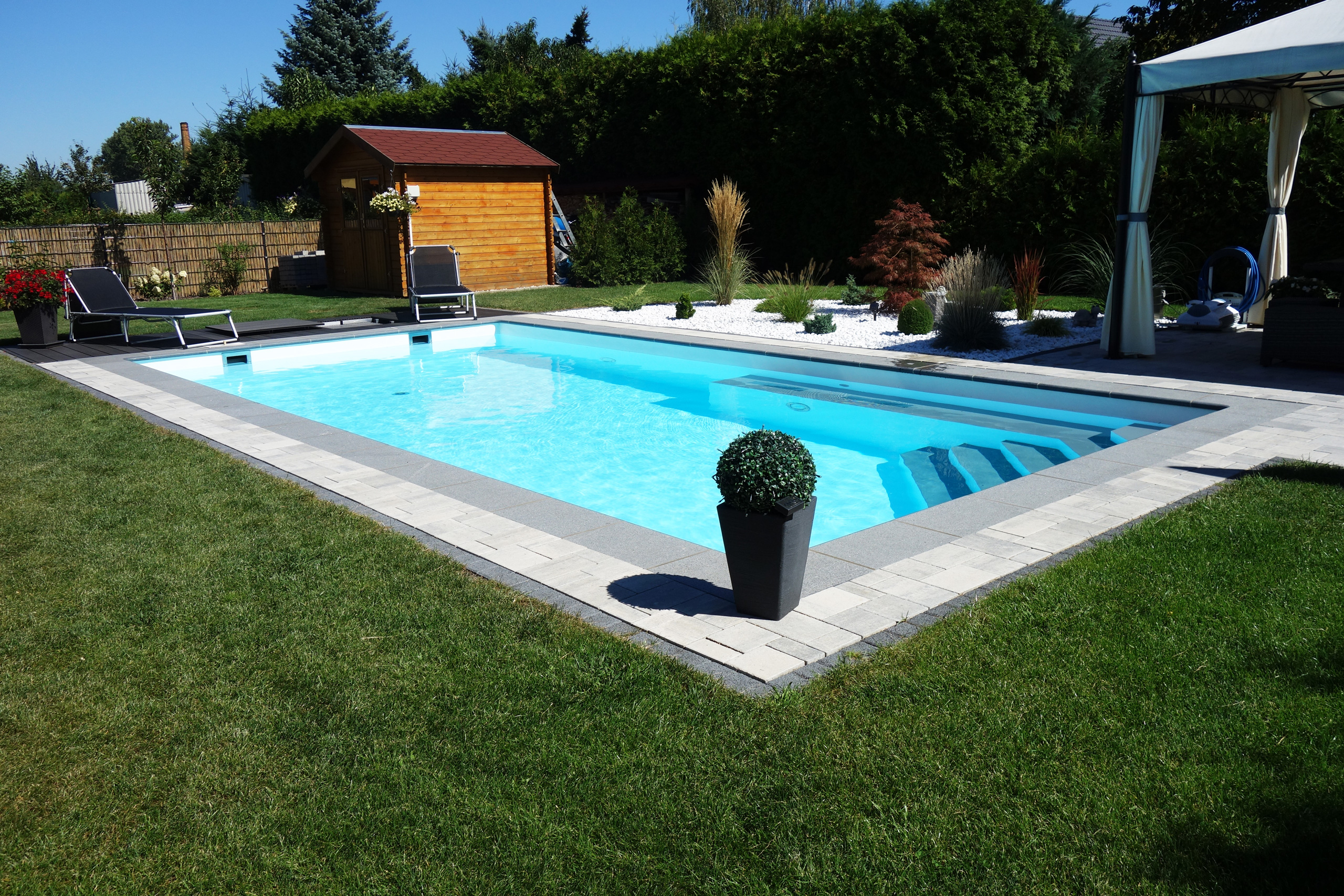Rechteckbecken mit Ecktreppe und Podest, umgeben von Rasen und einer gepflasterten Poolumrandung