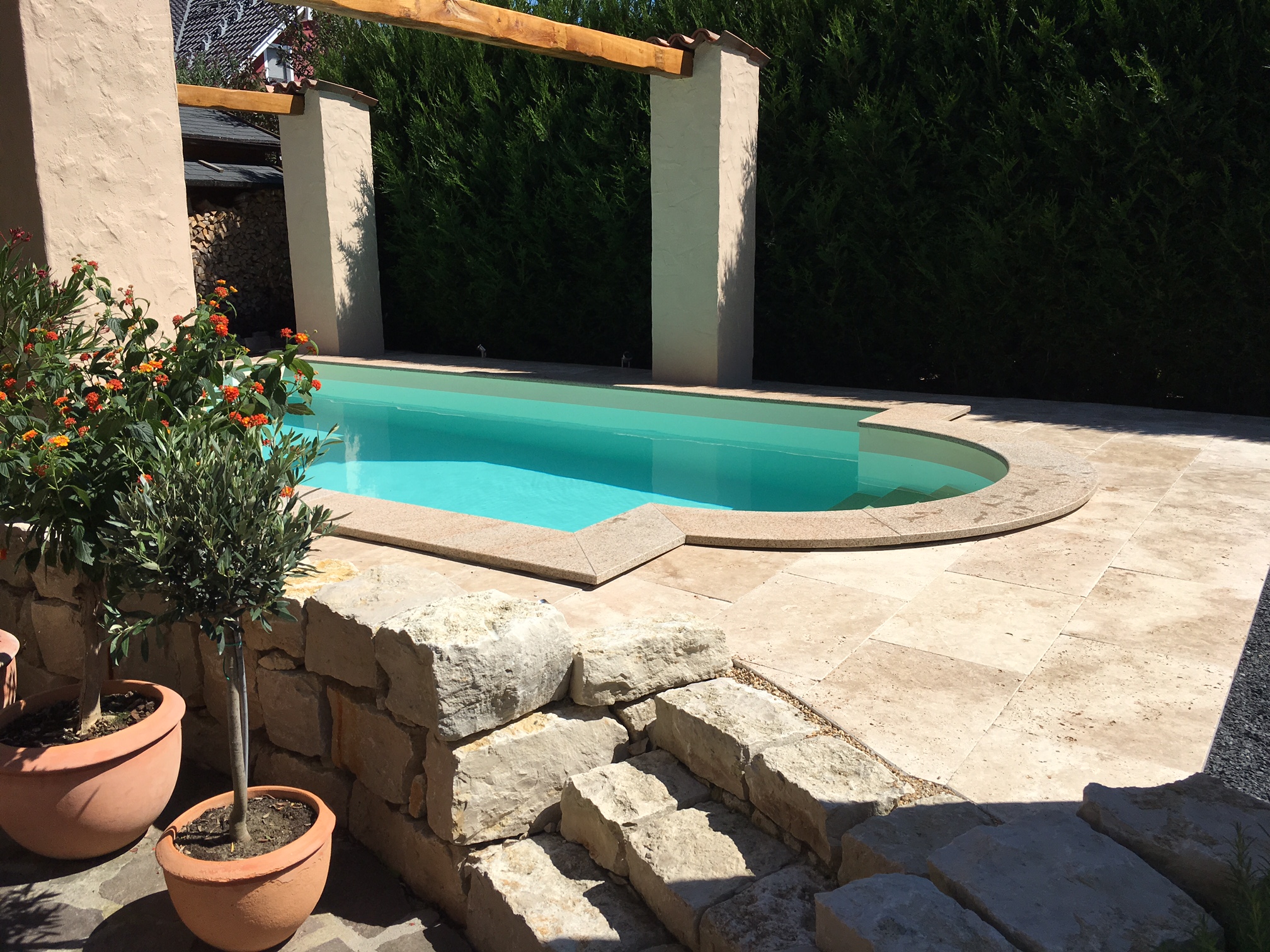 Schwimmbecken mit römischer Treppe, umgeben von hellen Steinplatten und einer grünen Hecke.