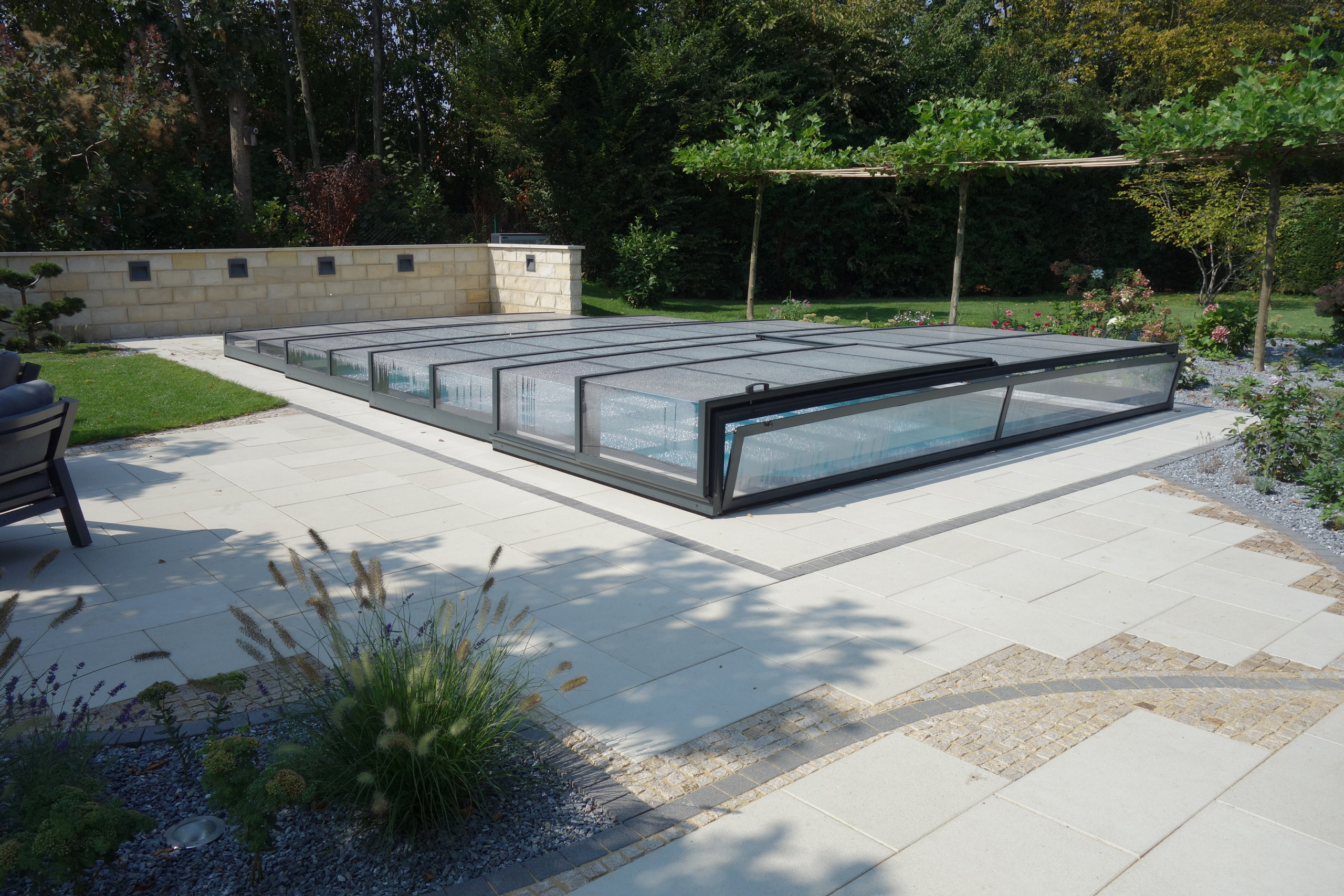 Überlaufbecken Overflow Stone mit geschlossener, flacher Poolüberdachung und heller Pflasterung im Garten