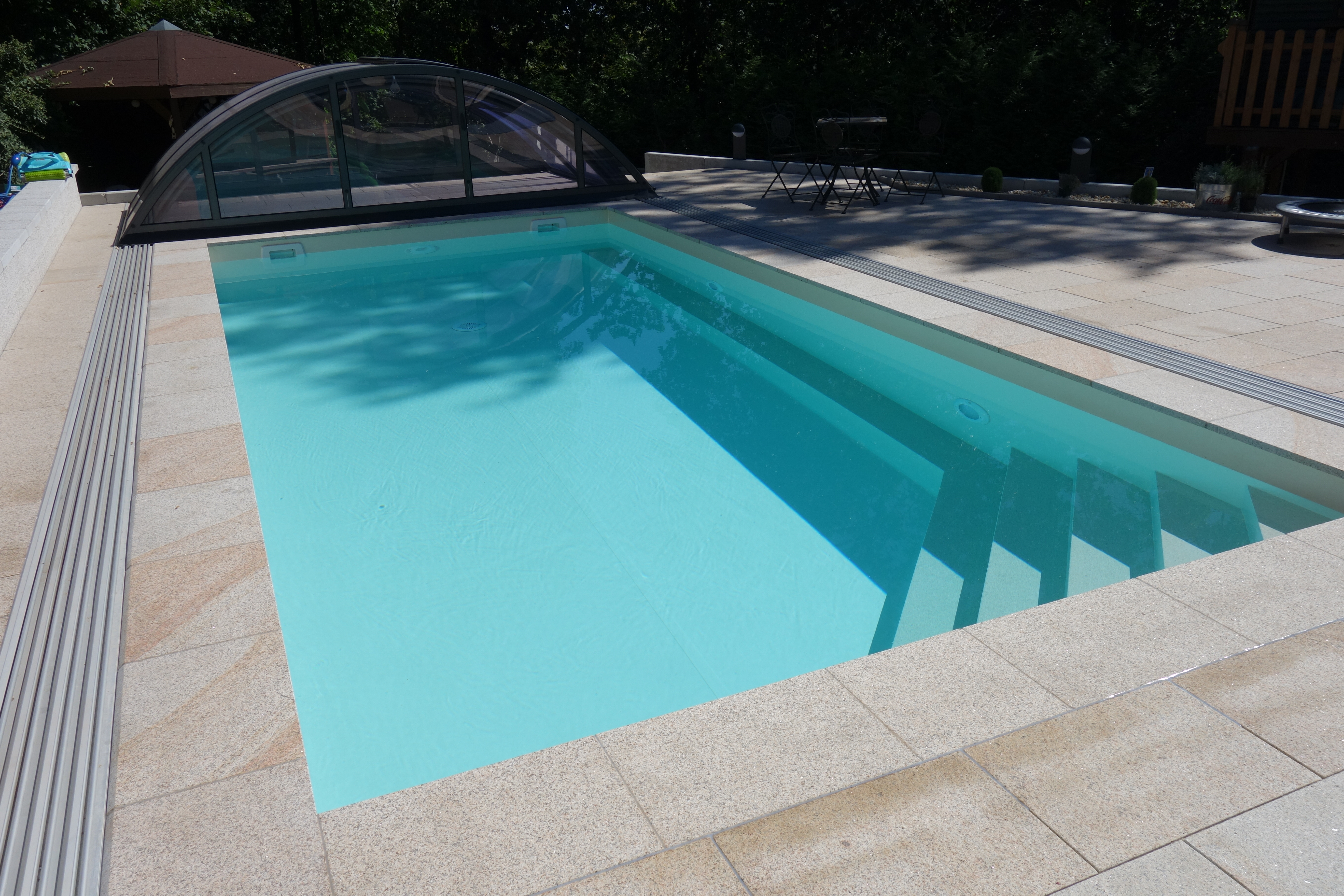 Rechteckpool mit Ecktreppe, Podest und Stehstufe, umgeben von hellen Platten und einer Überdachung