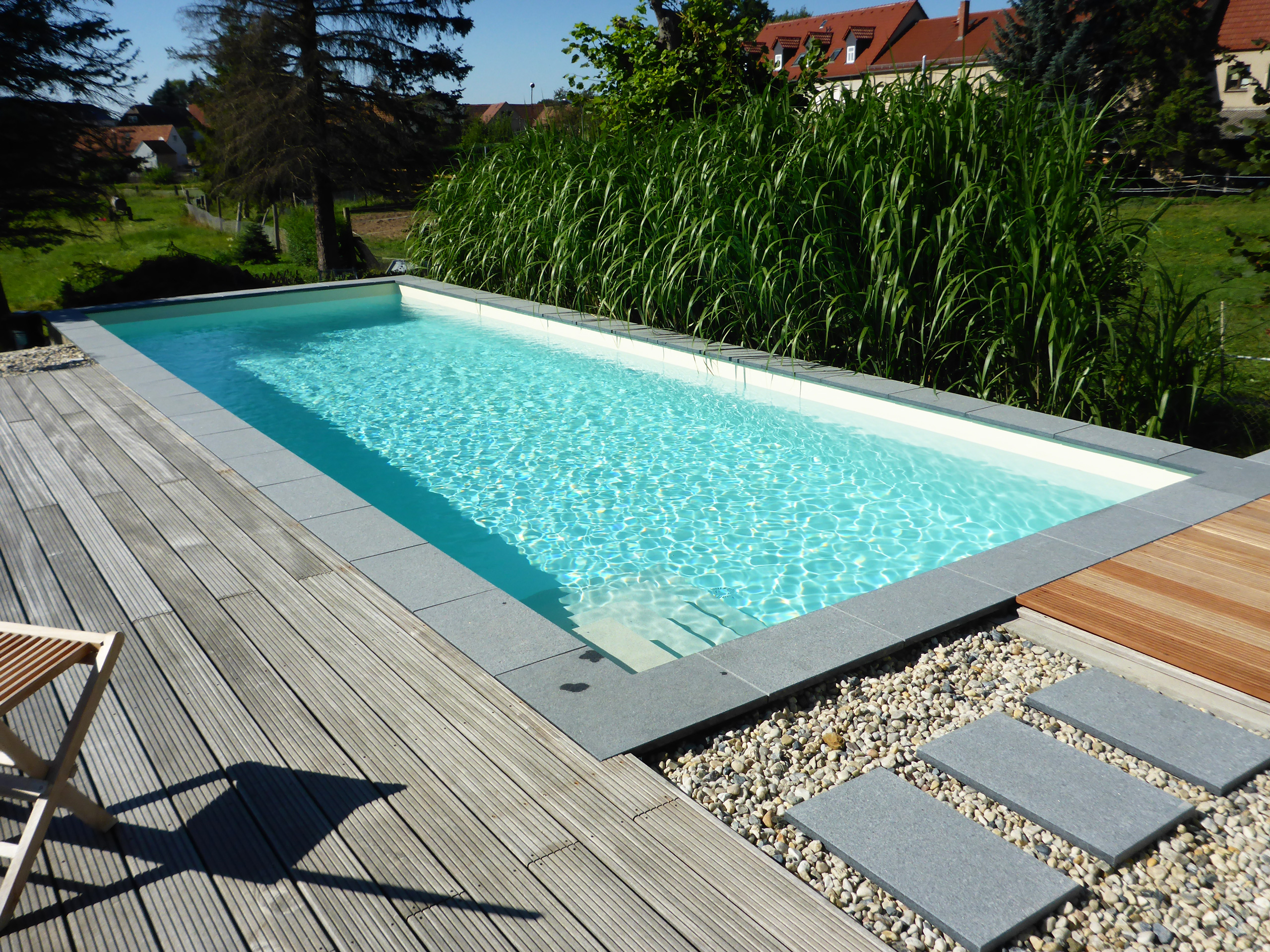 Rechteckiger Swimmingpool mit Ecktreppe, umgeben von Holzterrasse, Kies und Trittplatten.