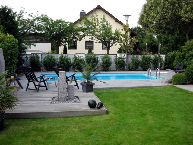Garten mit türkisem Schwimmbecken, Holzterrasse, Liegestühlen und einem Wasserspiel aus Stein.
