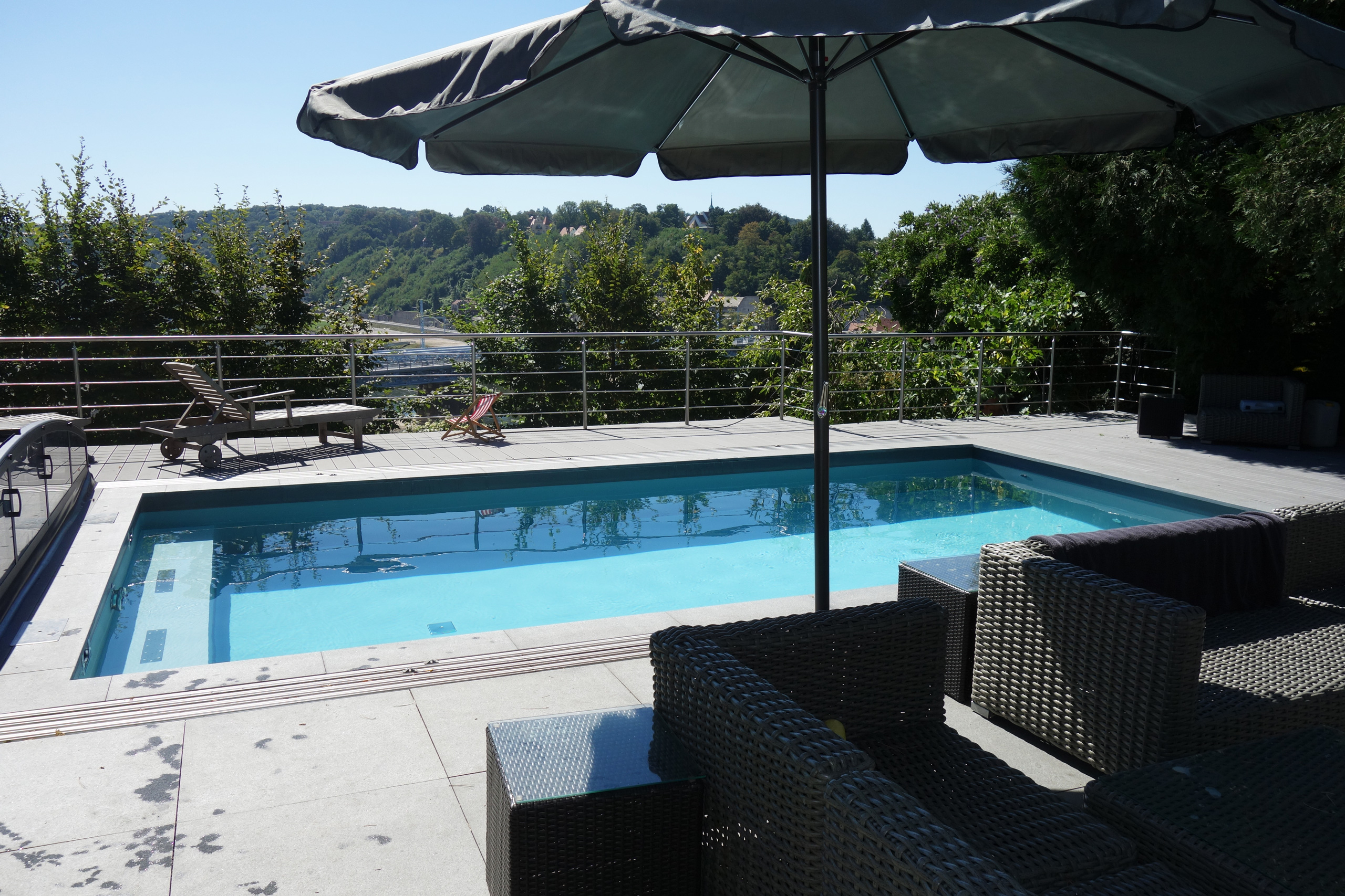 Blick auf das Betonbecken mit blauer Folie, Ecktreppe und Sitzbank, umgeben von einer Terrasse mit Möbeln.
