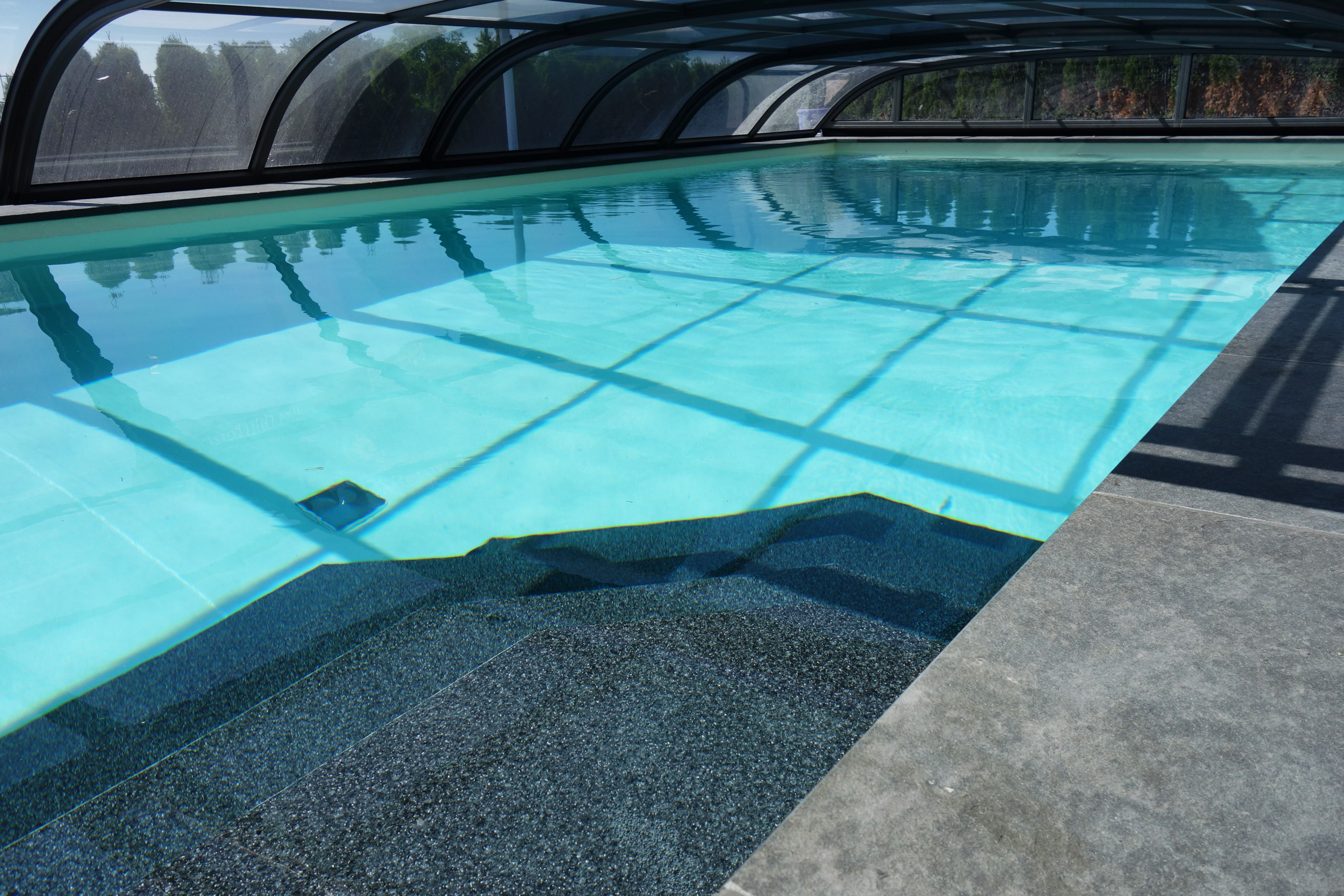 Treppe mit dunklem Granulat im hellblauen Schwimmbad unter einer transparenten Poolüberdachung