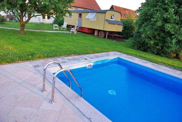 Rechteckbecken mit blauer Einhängefolie und Edelstahlleiter, umgeben von gepflasterter Poolumrandung und Rasen.