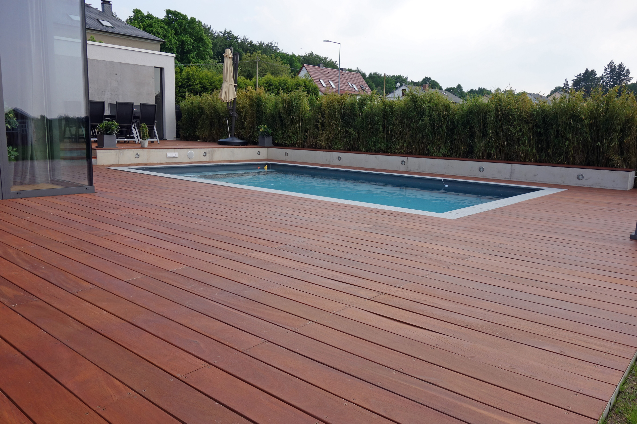 Betonbecken 3,5x7x1,5m mit Ecktreppe und Podest, umgeben von Holzterrasse und Bepflanzung