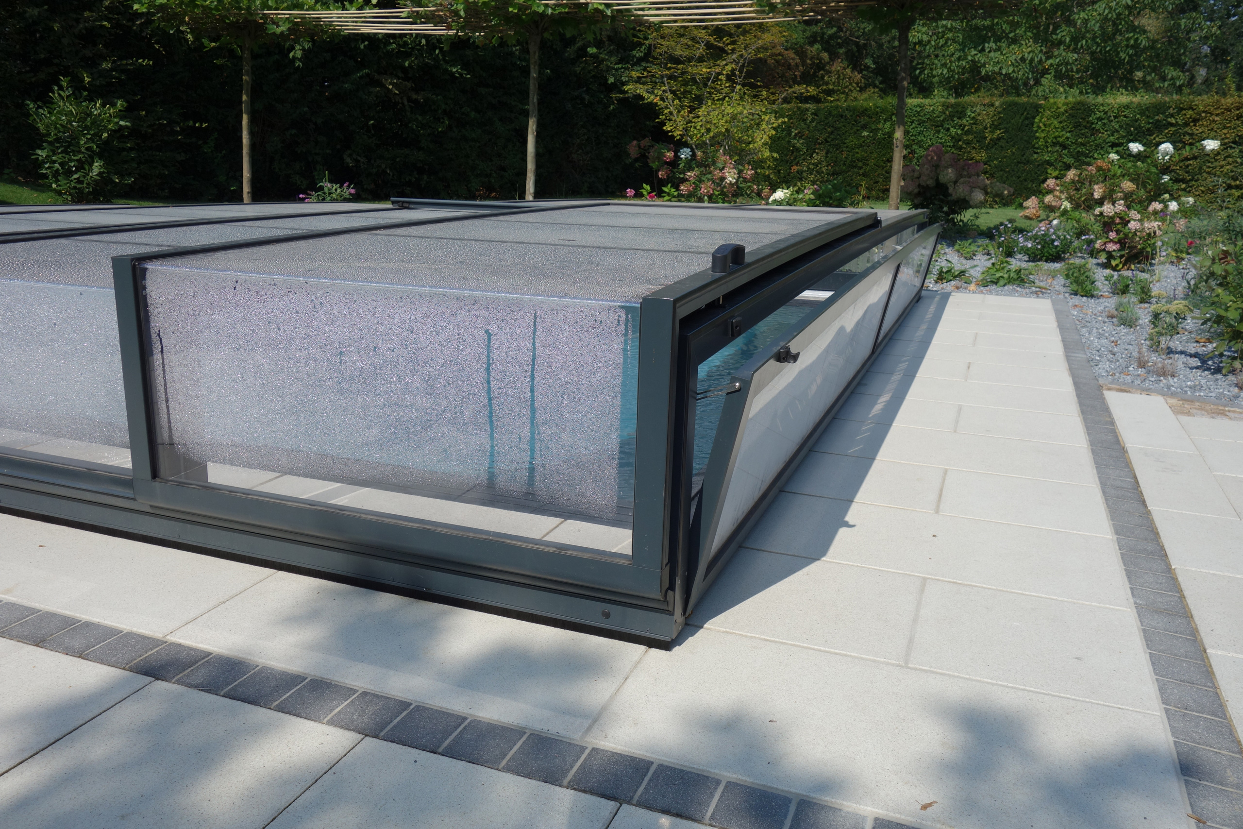 Detailansicht einer Poolabdeckung mit transparenten Elementen und dunklem Rahmen, auf hellen Terrassenplatten