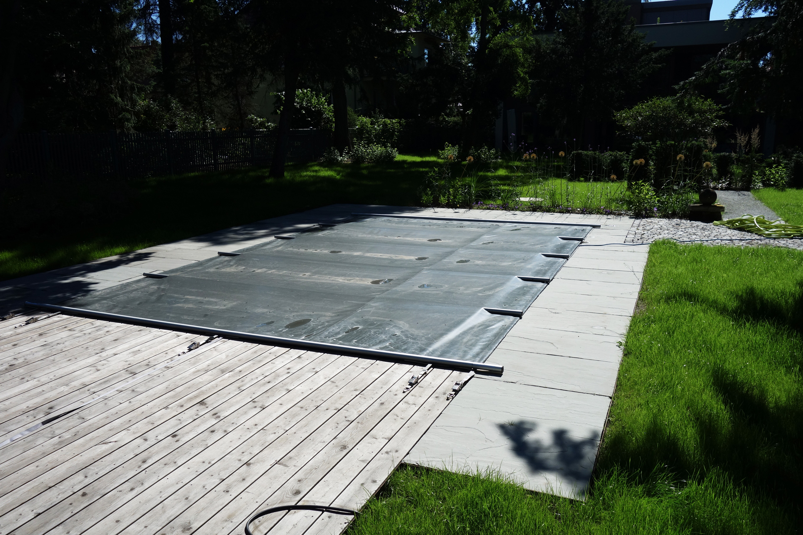 Abgedeckter Rechteck-Pool mit Holzterrasse und Steinplatten, umgeben von grünem Rasen und Bäumen.