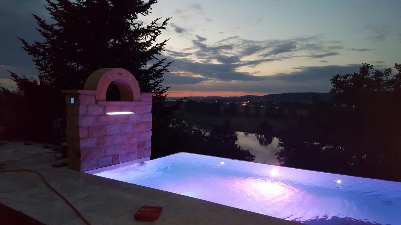 Überlaufbecken Infinity Pool mit beleuchtetem Wasserfall und Blick auf Flusslandschaft bei Sonnenuntergang
