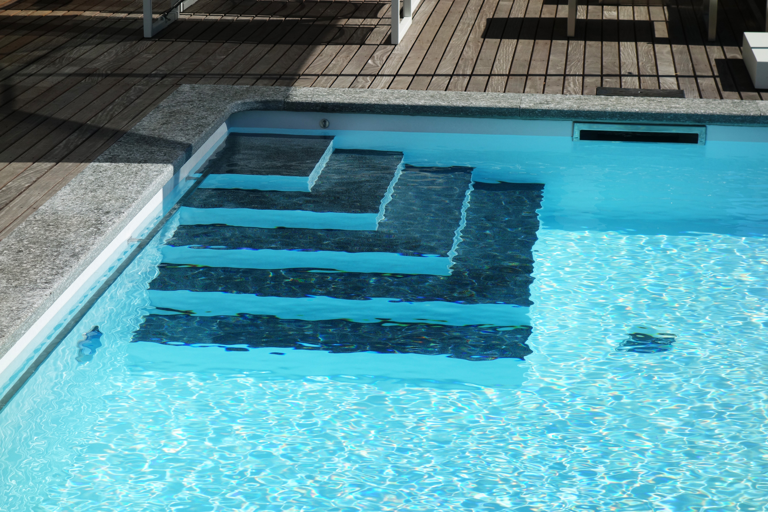 Ecktreppe und Podest eines Rechteckbeckens mit Unterflurabdeckung, umgeben von Holzterrasse und Steinrand.