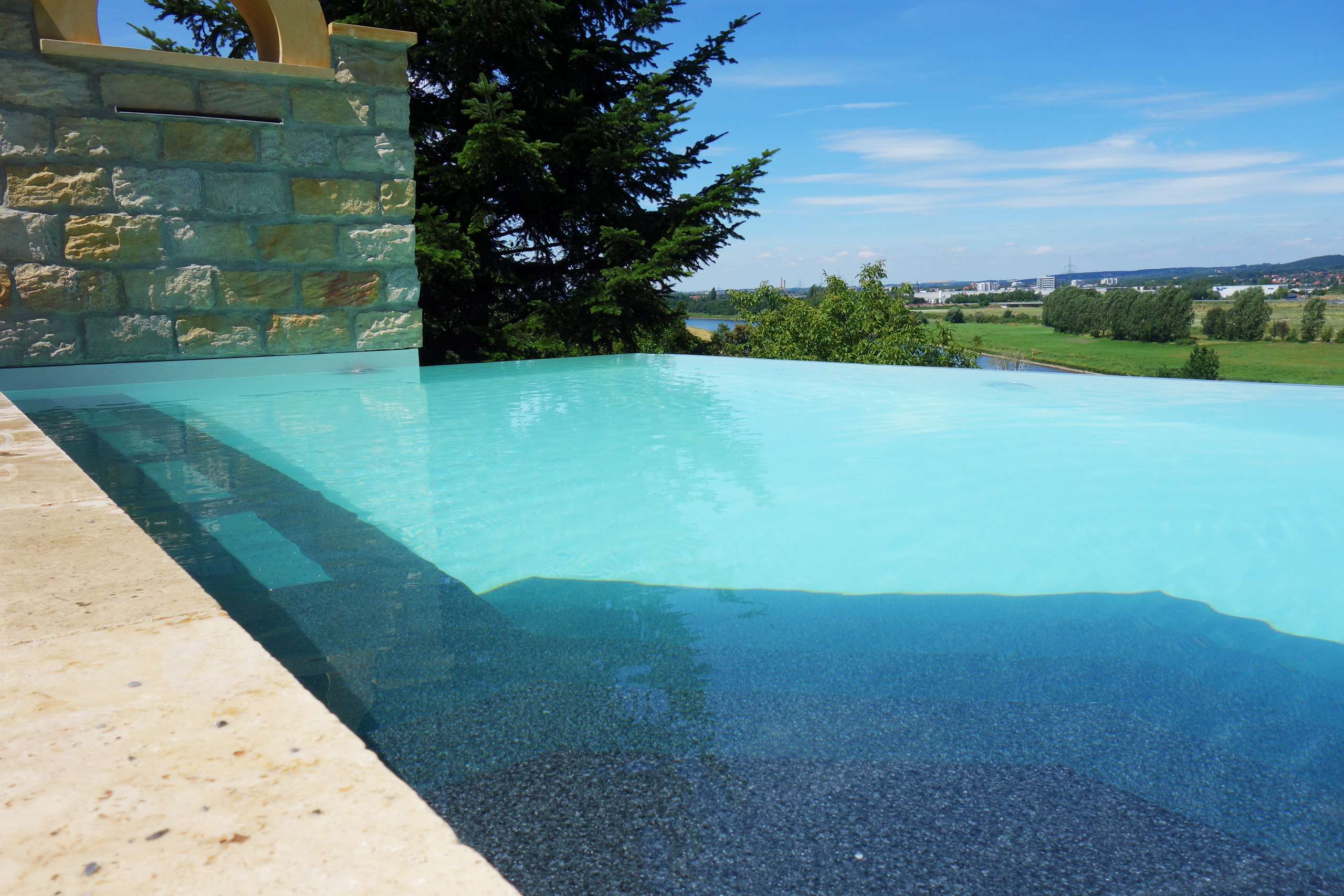 Blick über das Überlaufbecken mit hellblauem Wasser und dunklem Boden auf eine weite Landschaft