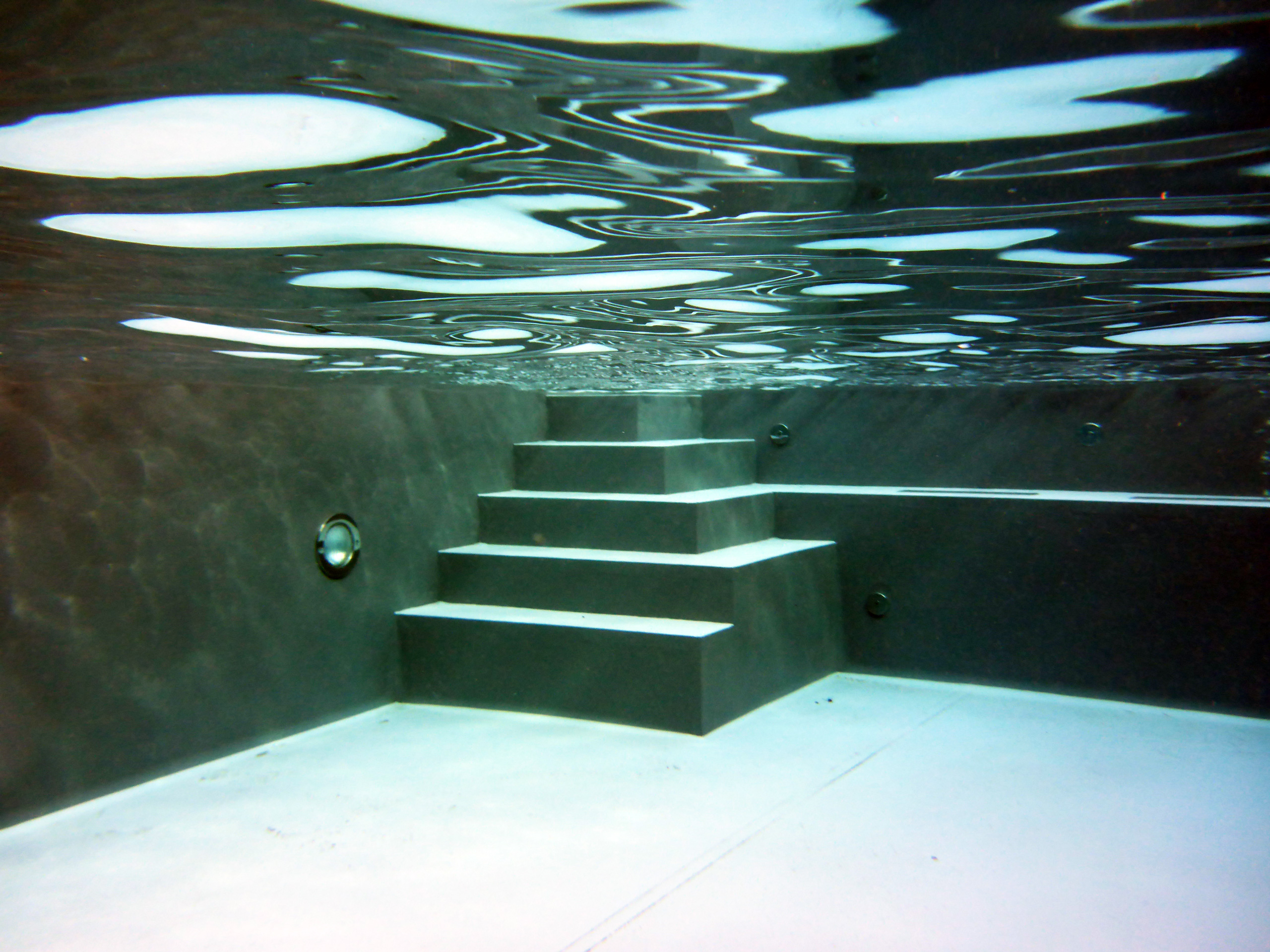 Unterwasseransicht eines Betonbeckens mit Ecktreppe und Podest, die Wasseroberfläche spiegelt sich.