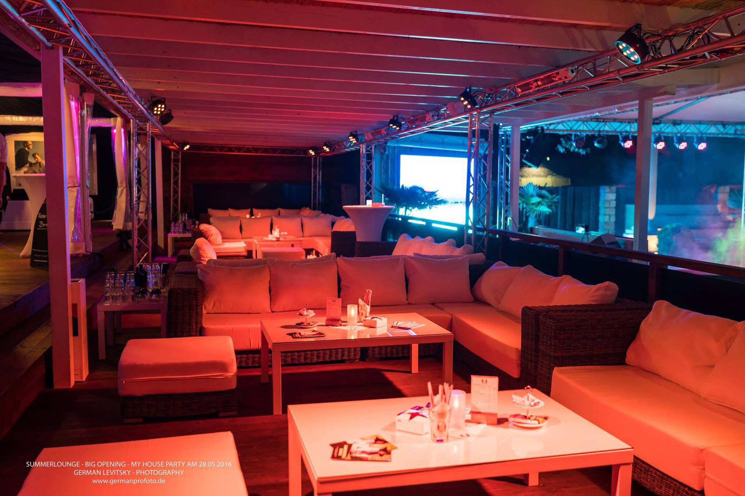 Beleuchtete Lounge mit Rattan-Sofas und Tischen, im Hintergrund ein Pool mit blauer Beleuchtung.