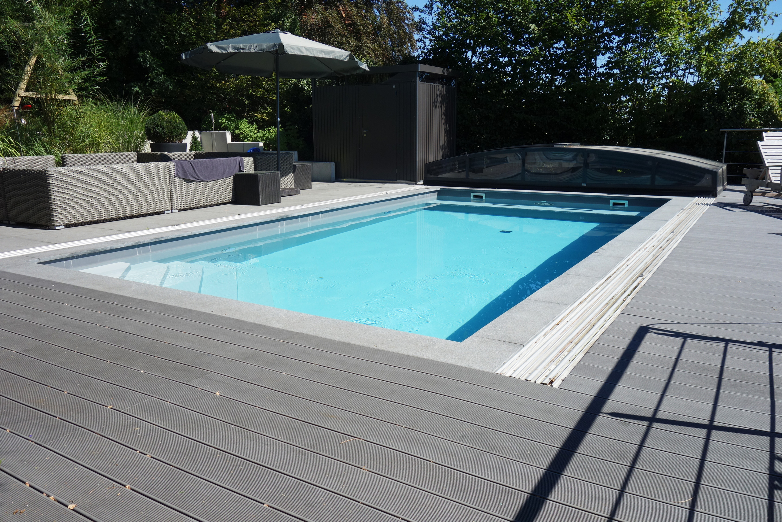 Betonbecken mit Ecktreppe und Sitzbank, umgeben von dunklem Holzdeck und einer Überdachung