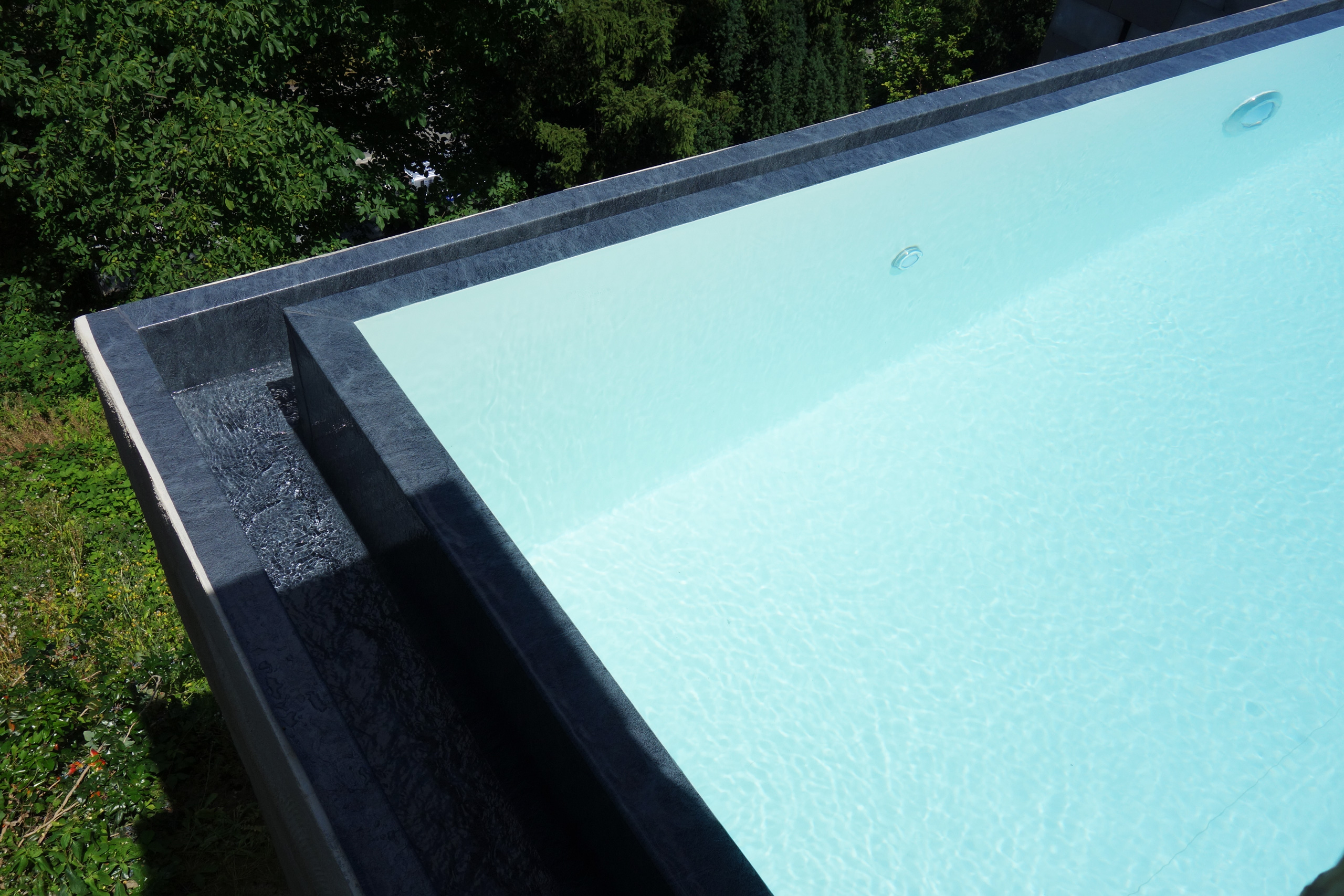 Blick von oben auf den Überlaufkanal eines Infinity Pools mit hellblauem Wasser und grüner Umgebung.
