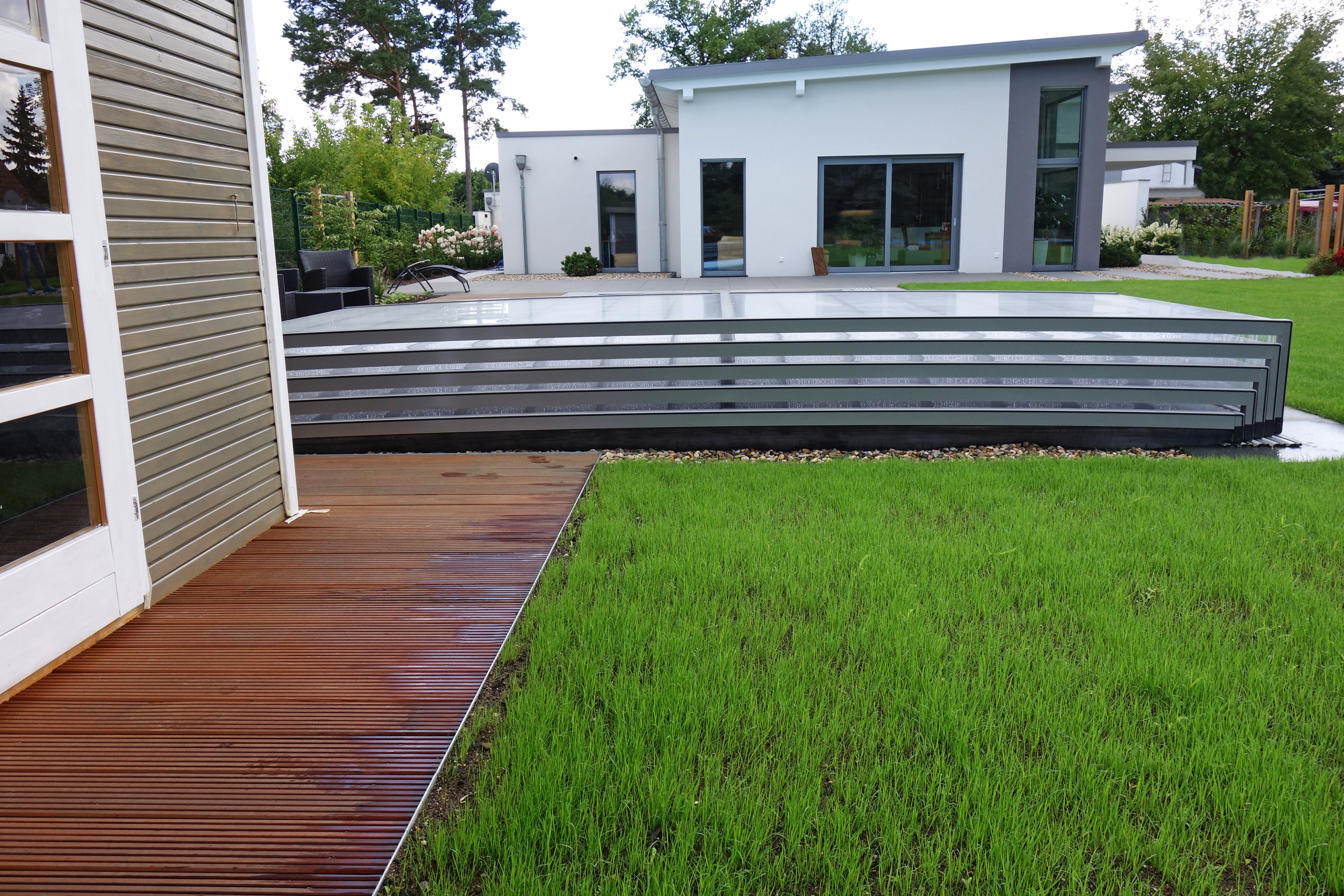 Blick auf den Außenpool mit Überdachung, Holzterrasse und Rasenfläche im Garten eines modernen Hauses.