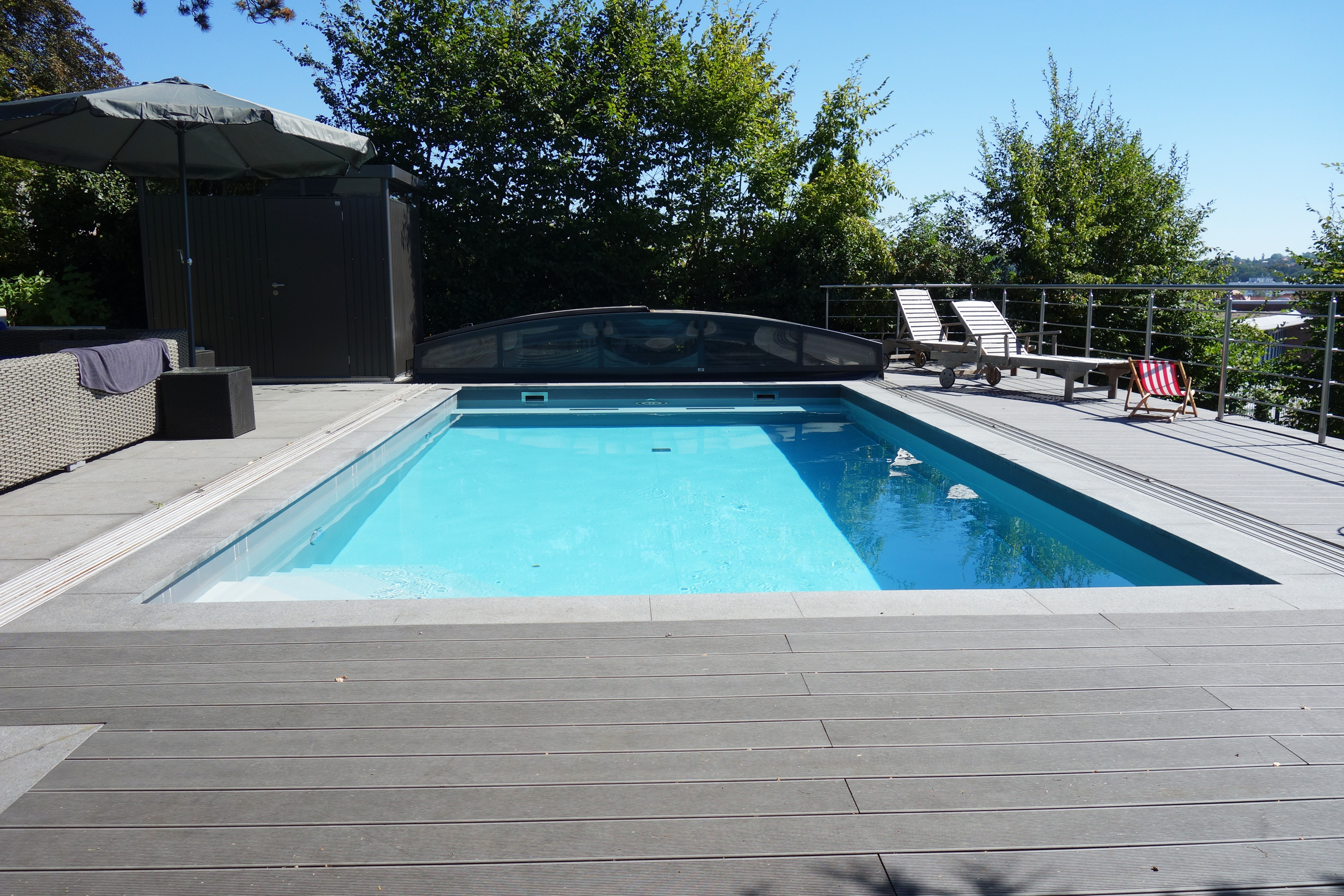 Betonbecken mit Überdachung, umgeben von einer dunklen Holzterrasse und einem Sonnenschirm